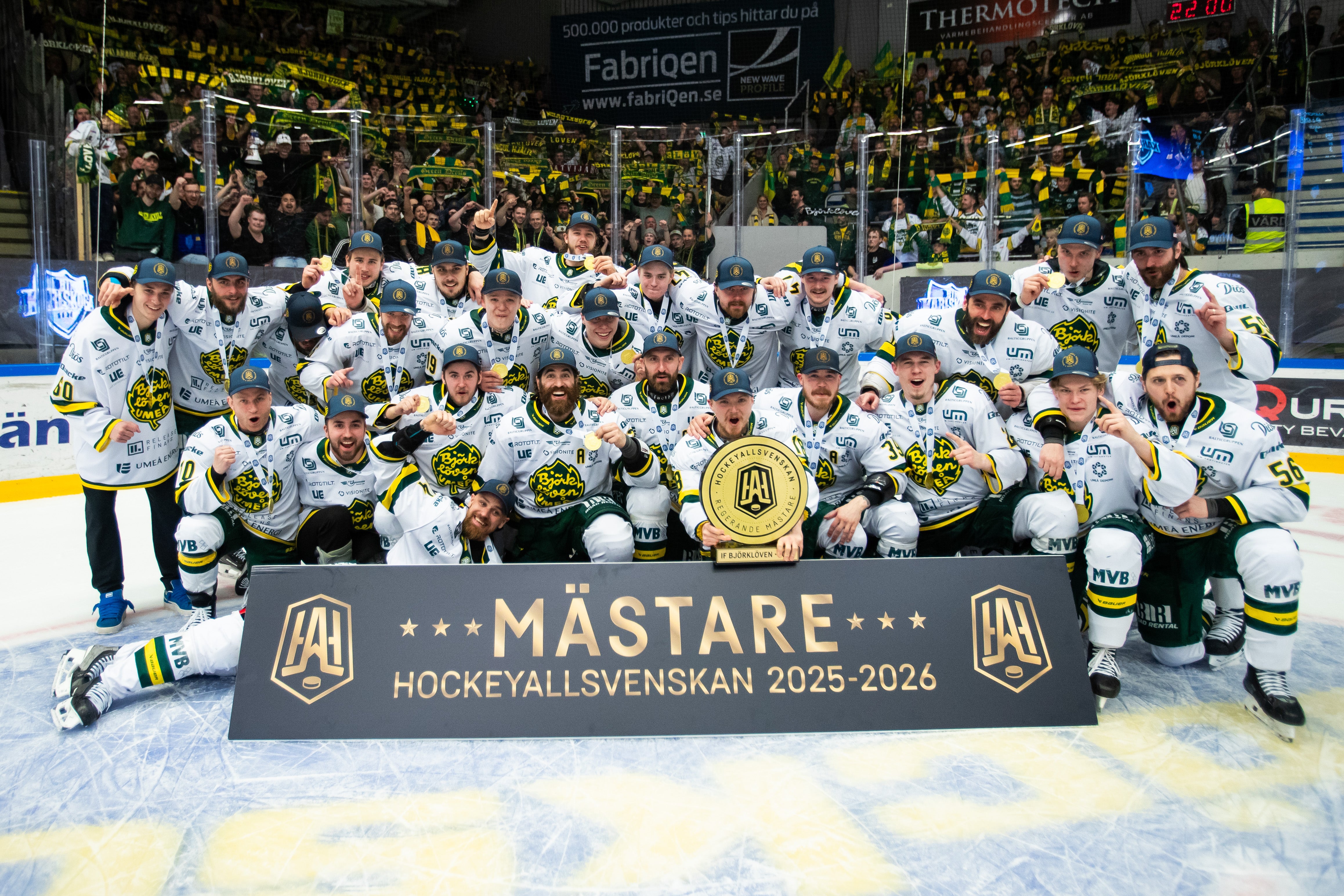 Ett ishockeylag i vita tröjor poserar tillsammans på isen med en stor guldpokal och en mästarbanner efter att ha vunnit Hockeyallsvenskan 2025-2026.