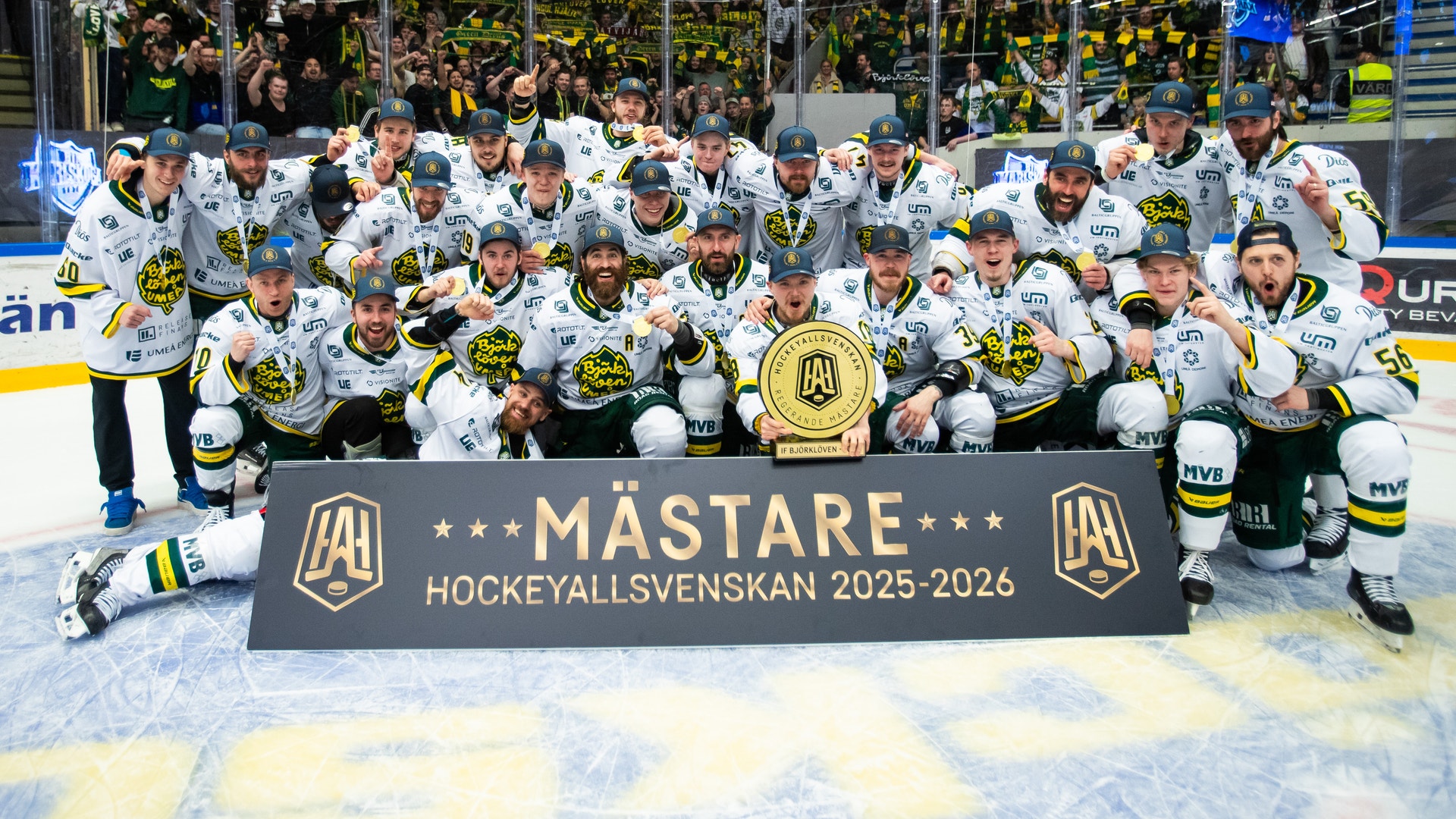 Ett ishockeylag i vita tröjor poserar tillsammans på isen med en stor guldpokal och en mästarbanner efter att ha vunnit Hockeyallsvenskan 2025-2026.