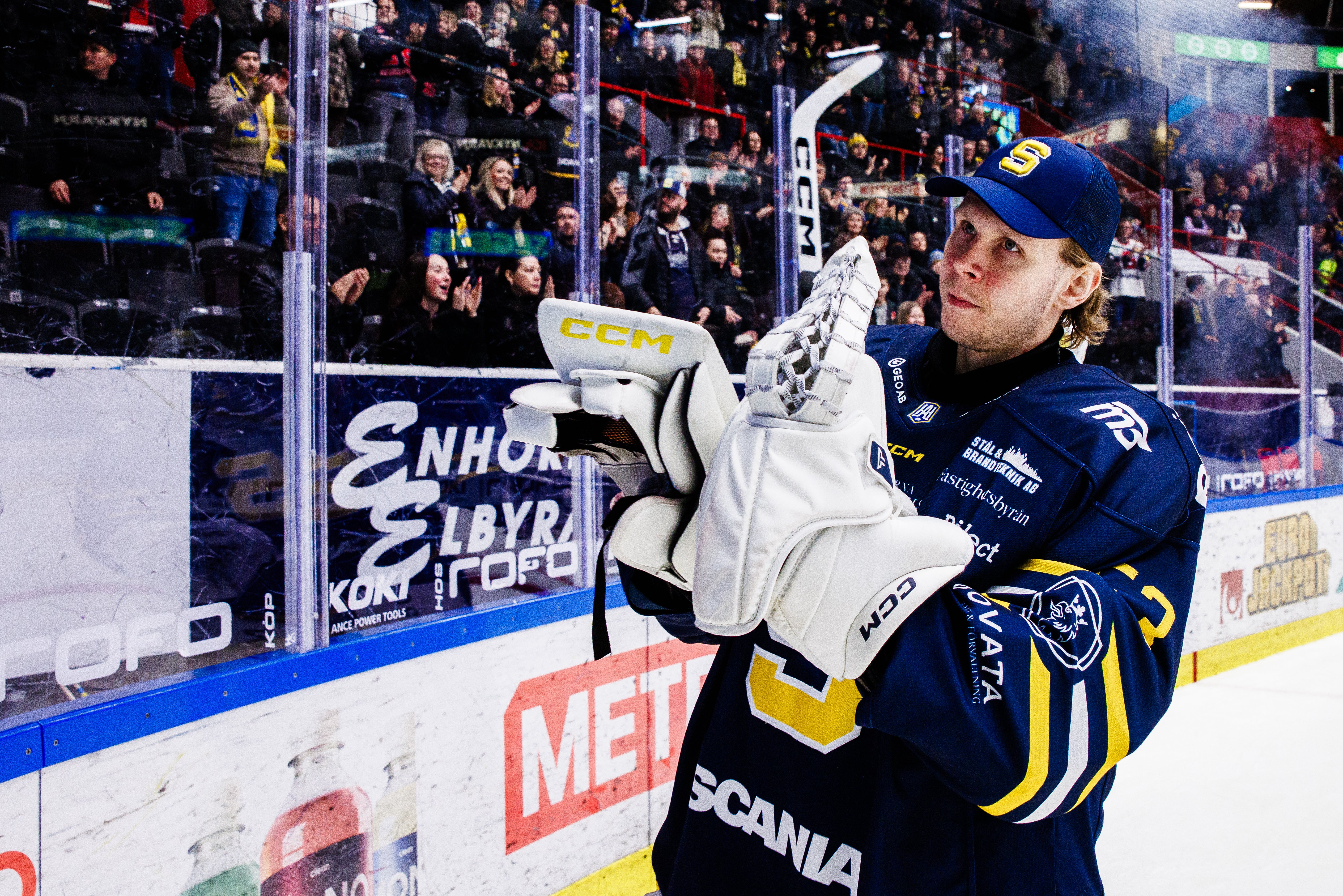 Ishockeyspelare i mörkblå tröja slår puck mot mål under en match. Spelaren bär blå keps och vita handskar. Åskådare och reklamskyltar syns i bakgrunden.
