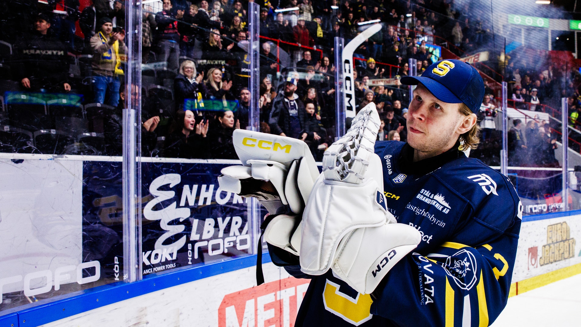 Ishockeyspelare i mörkblå tröja slår puck mot mål under en match. Spelaren bär blå keps och vita handskar. Åskådare och reklamskyltar syns i bakgrunden.