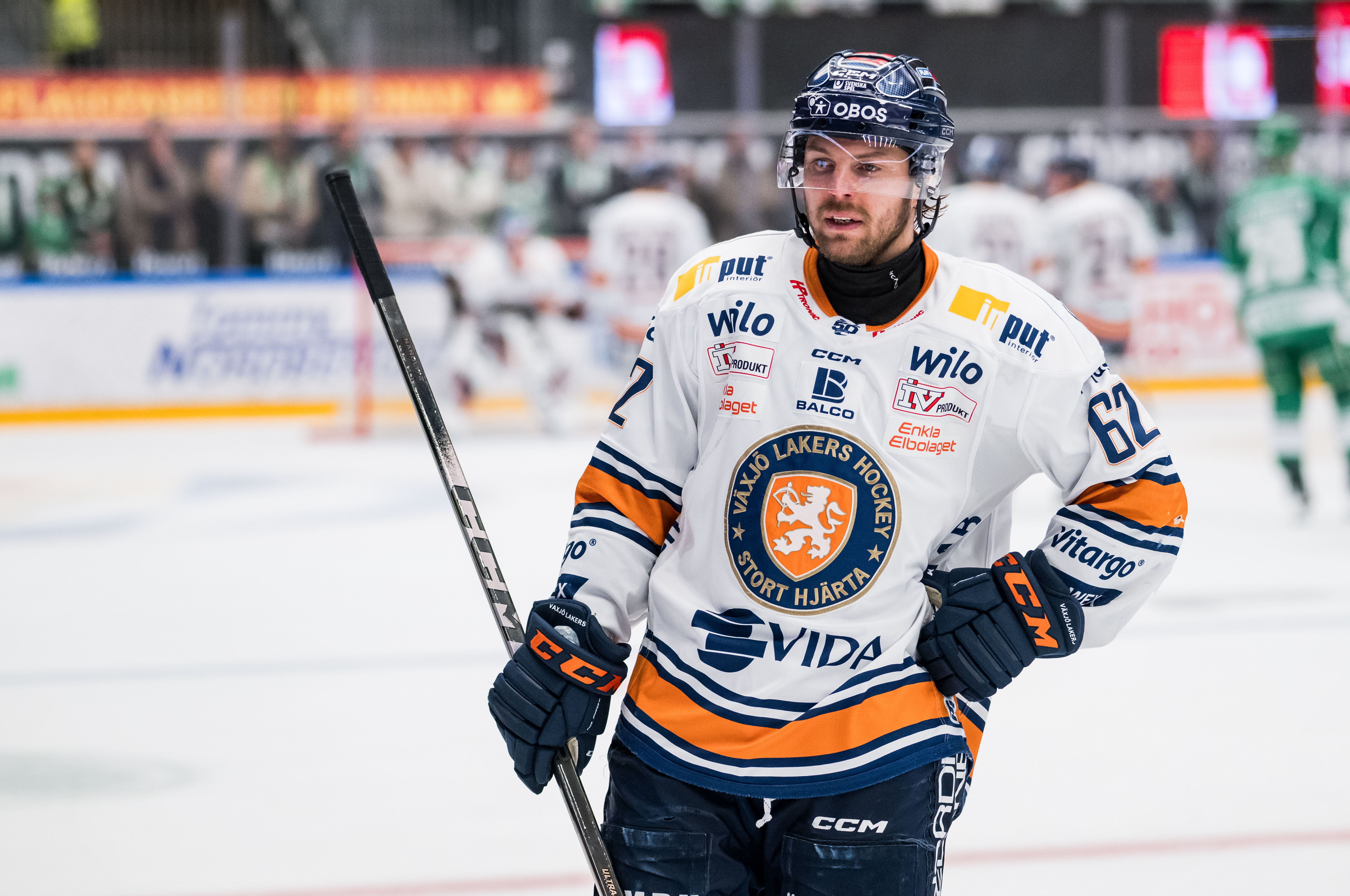 Ishockeyspelare i vit tröja med orange och mörkblå detaljer står på isen med sin hockeystick under en match.