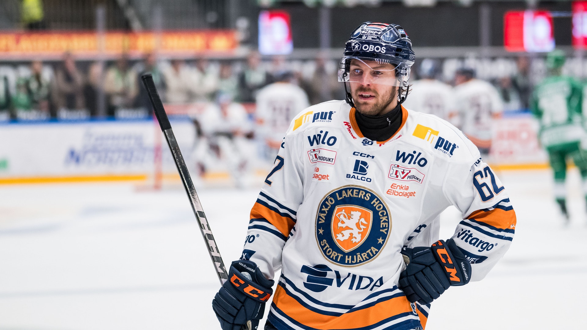 Ishockeyspelare i vit tröja med orange och mörkblå detaljer står på isen med sin hockeystick under en match.