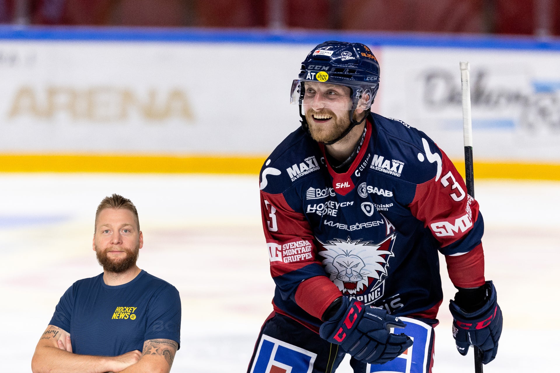 Ishockeyspelare i mörkblå och röd tröja på isen tillsammans med en man i marinblå t-shirt.