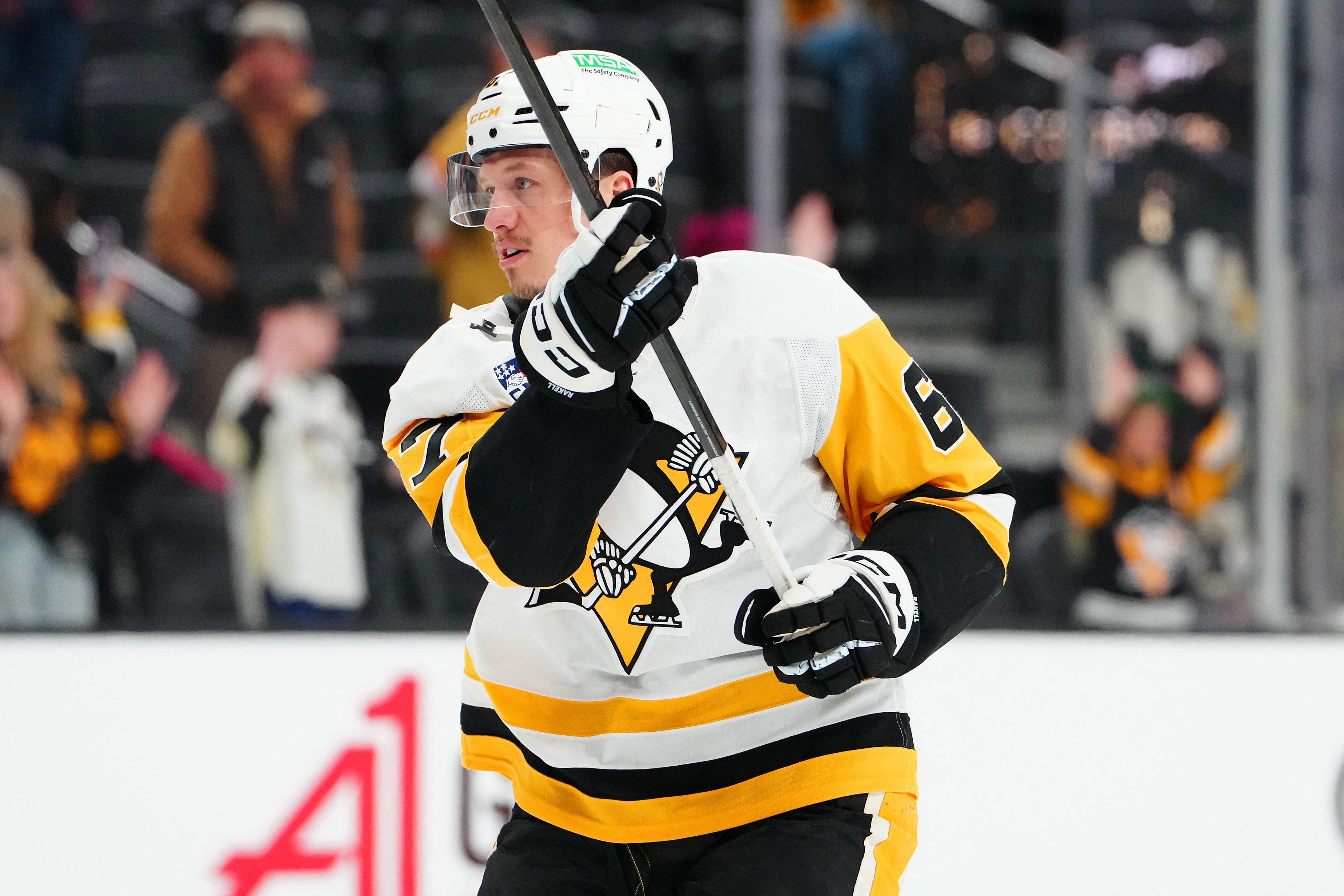 Ishockeyspelare i Pittsburgh Penguins svart och gul uniform håller i sin hockey stick under en match. Spelaren bär hjälm och handskar. Publiken syns suddig i bakgrunden.