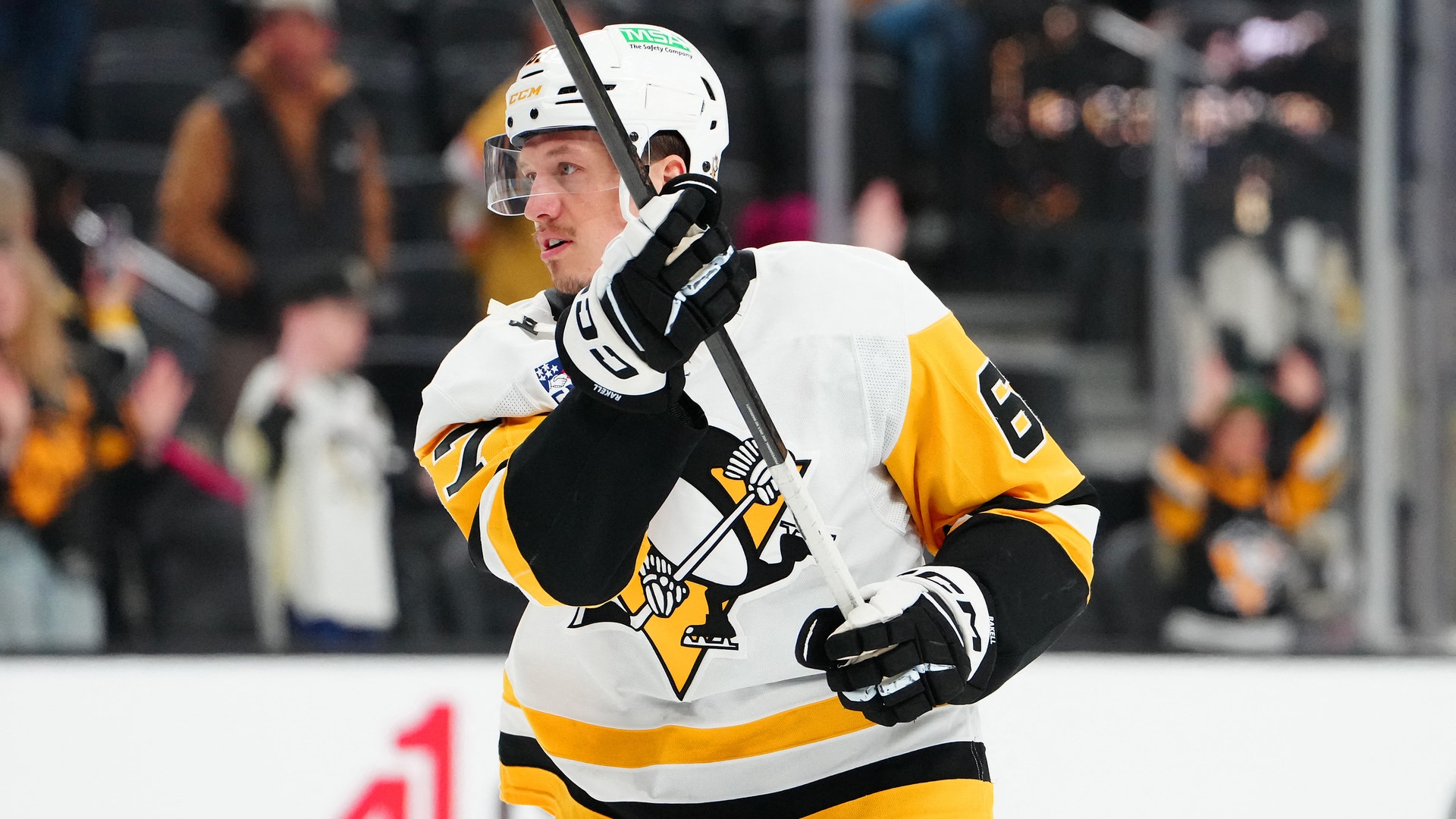 Ishockeyspelare i Pittsburgh Penguins svart och gul uniform håller i sin hockey stick under en match. Spelaren bär hjälm och handskar. Publiken syns suddig i bakgrunden.