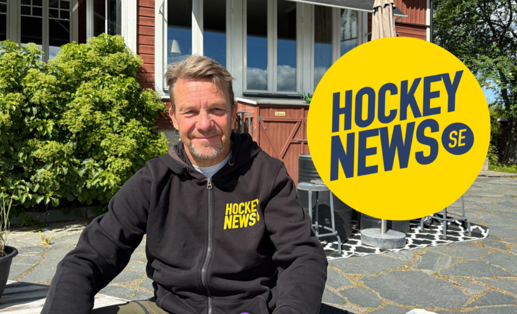 Porträttbild av en man i svart hoodie med Hockey News-logotyp, sittande utomhus framför en byggnad med gröna växter i bakgrunden.