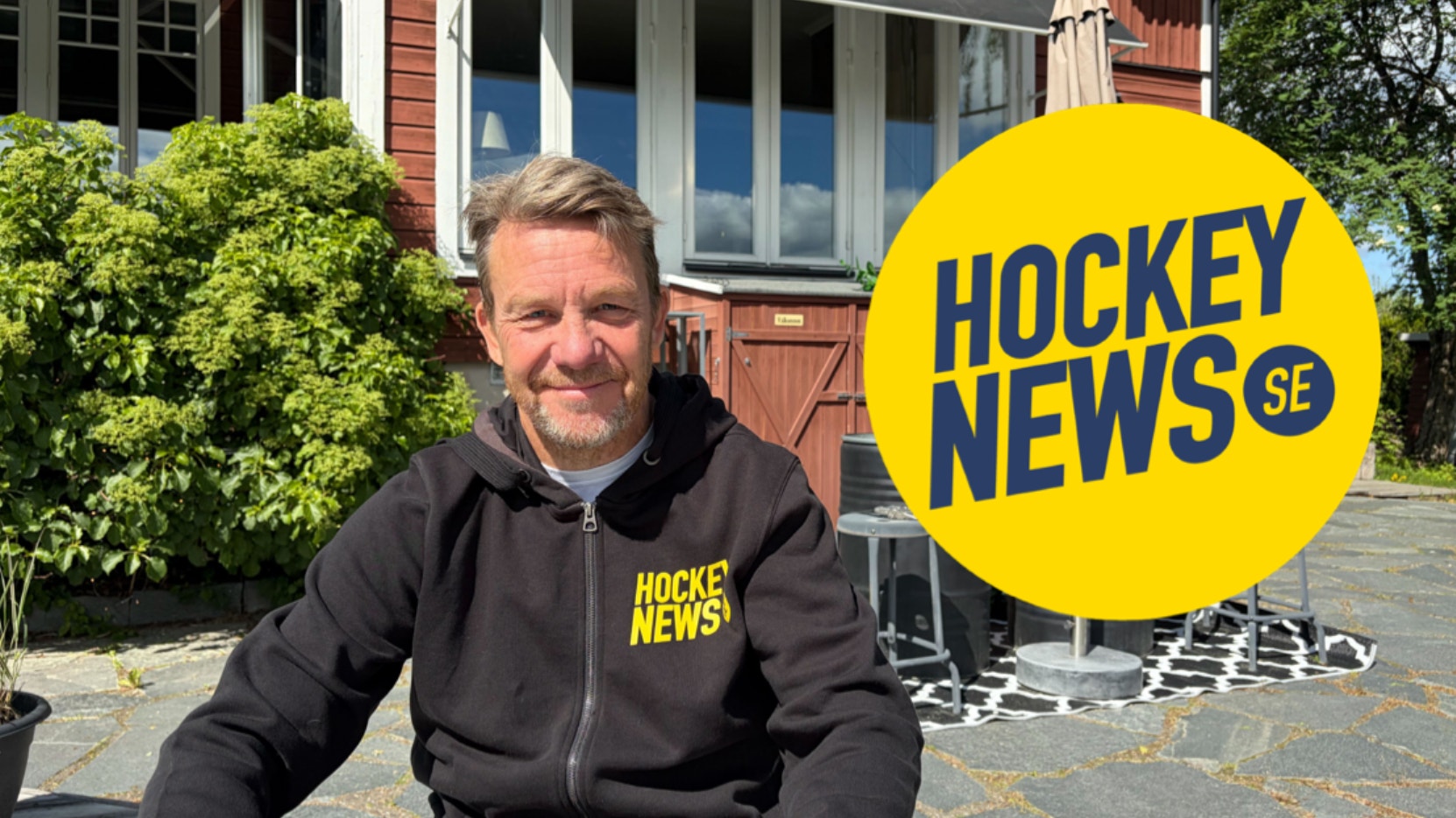 Porträttbild av en man i svart hoodie med Hockey News-logotyp, sittande utomhus framför en byggnad med gröna växter i bakgrunden.