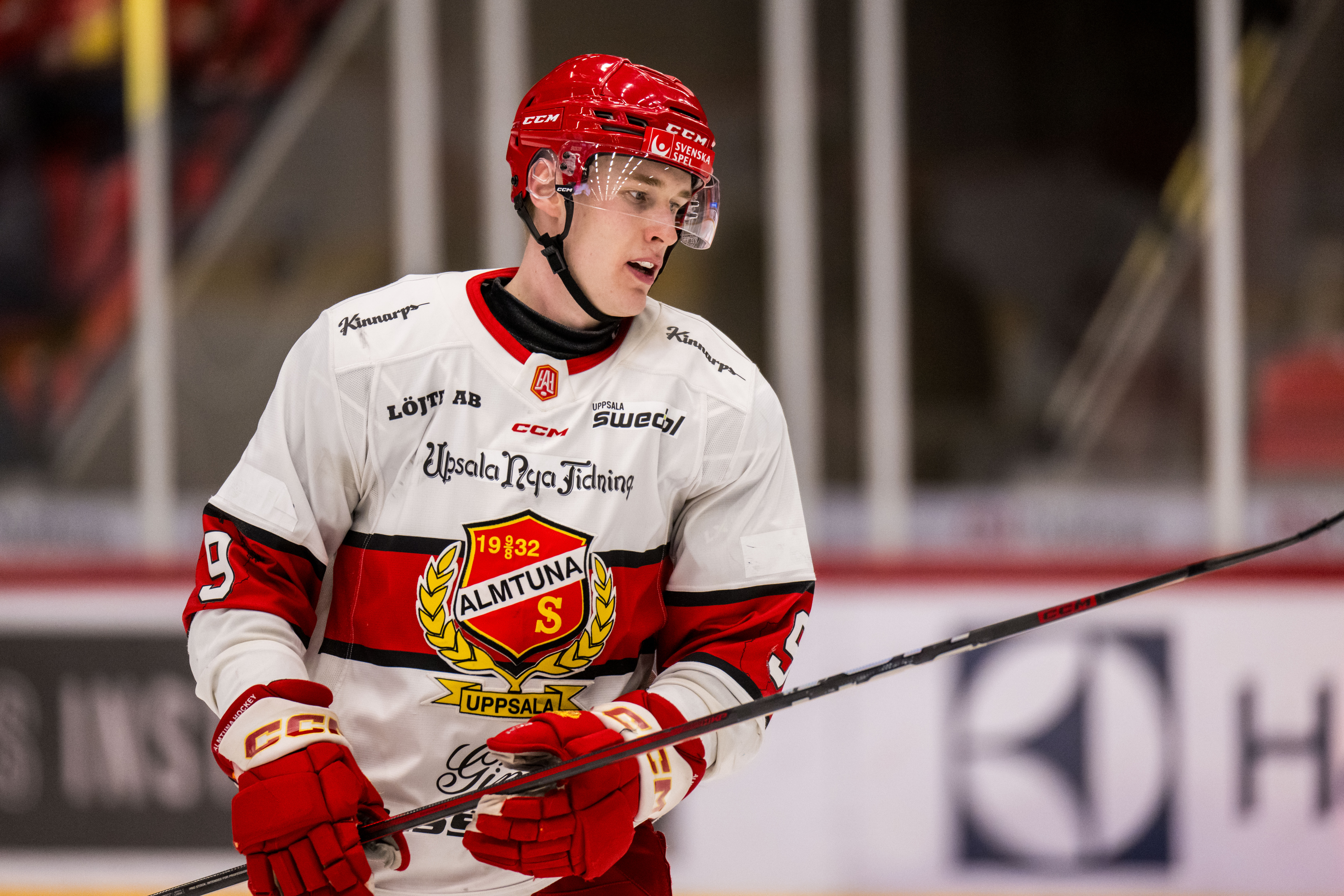 En ishockeyspelare i röd och vit dräkt med röd hjälm håller en ishockeyklubba under en match.