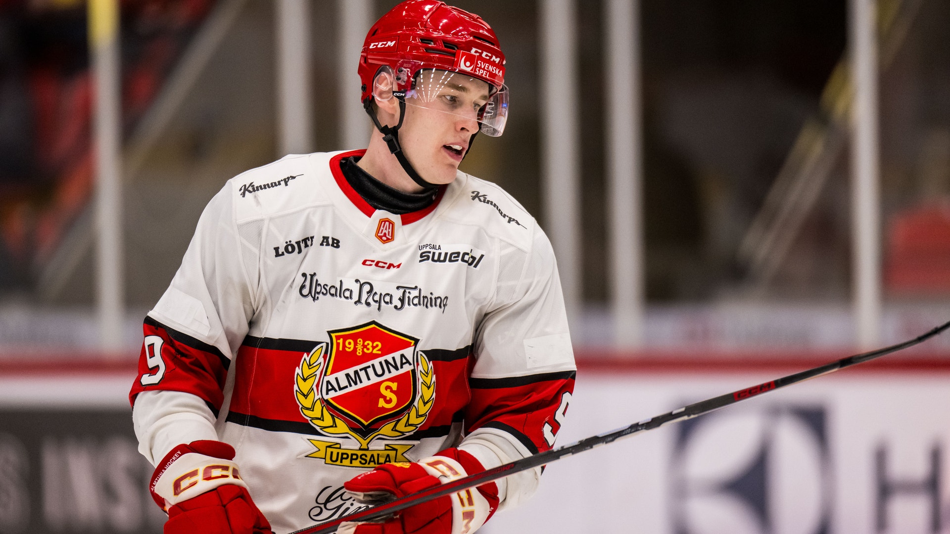 En ishockeyspelare i röd och vit dräkt med röd hjälm håller en ishockeyklubba under en match.