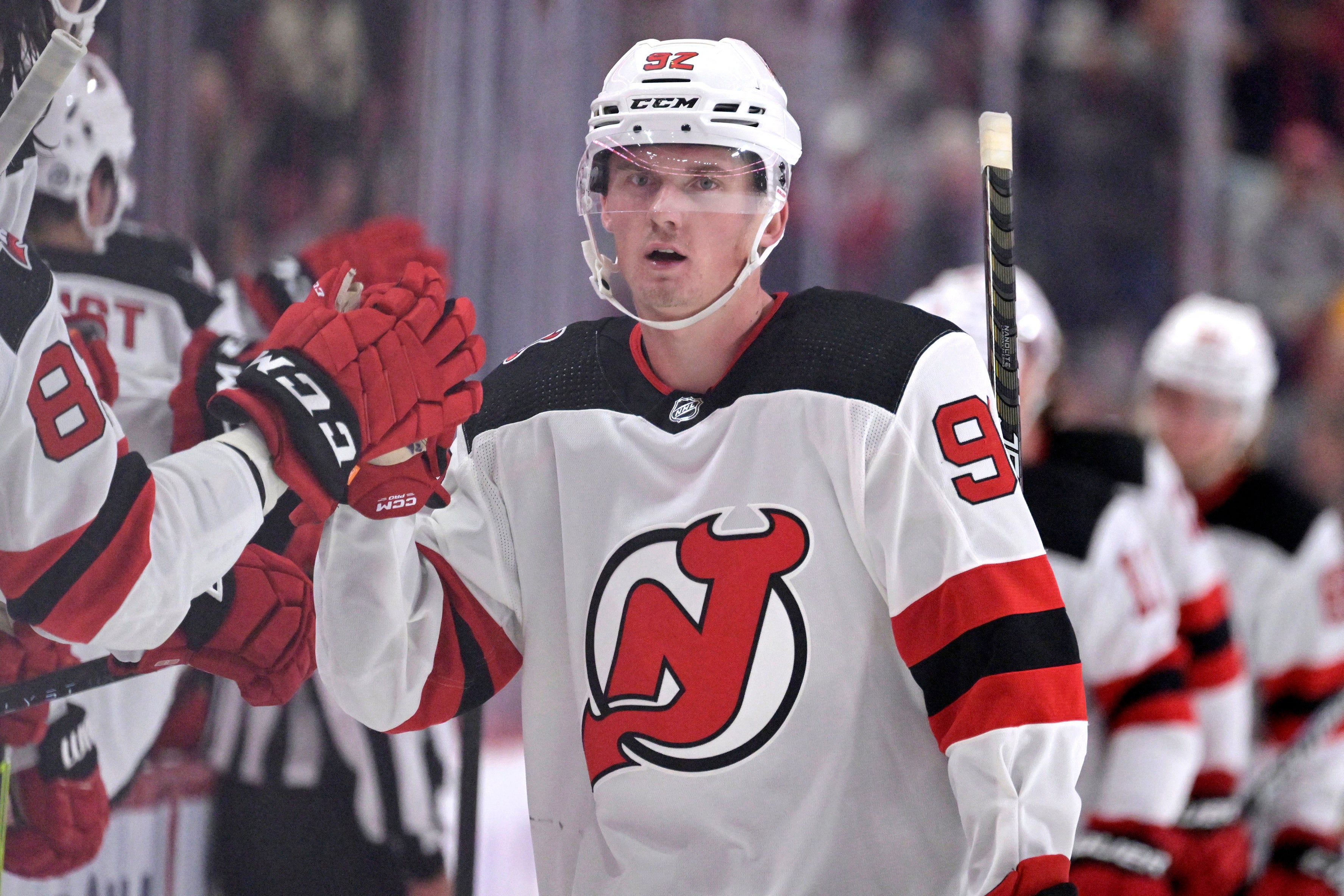 Ishockeyspelare i New Jersey Devils vita tröja med röda och svarta detaljer firar under en match. Spelaren bär vit hjälm och röda handskar, och höjer armen i glädje med publiken suddig i bakgrunden.