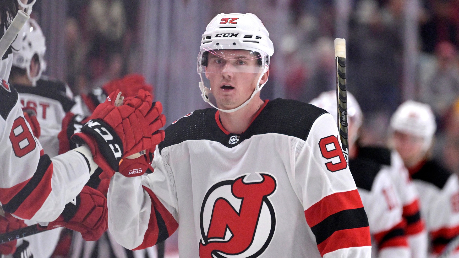 Ishockeyspelare i New Jersey Devils vita tröja med röda och svarta detaljer firar under en match. Spelaren bär vit hjälm och röda handskar, och höjer armen i glädje med publiken suddig i bakgrunden.