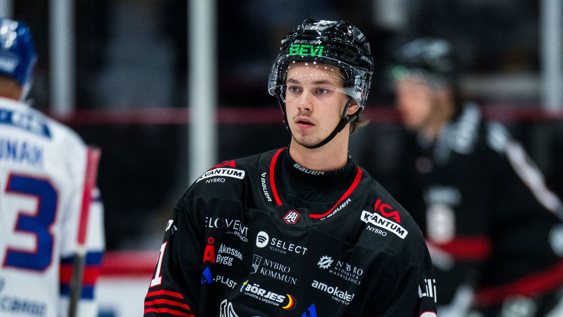 En ishockeyspelare i svart matchtröja och svart hjälm med visir står på isen i en hall. I bakgrunden syns suddigt andra spelare.