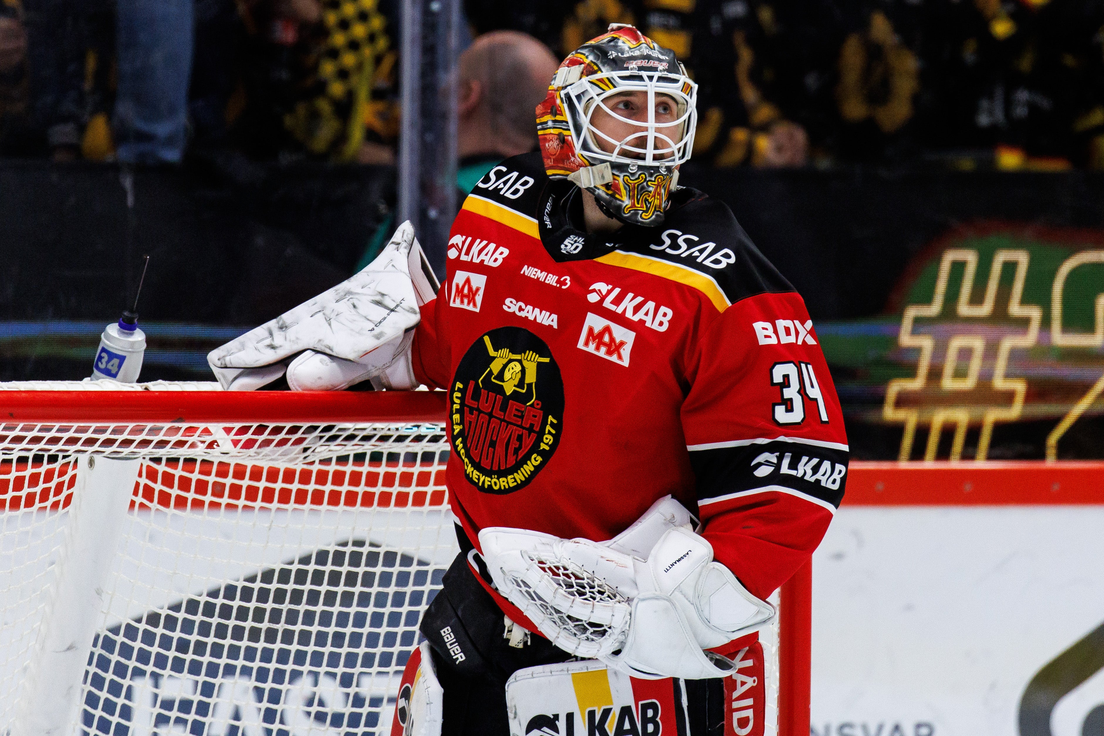 Ishockeyspelare i vit, gul och grön matchdräkt på isen under en match. Andra spelare och publik syns suddig i bakgrunden.