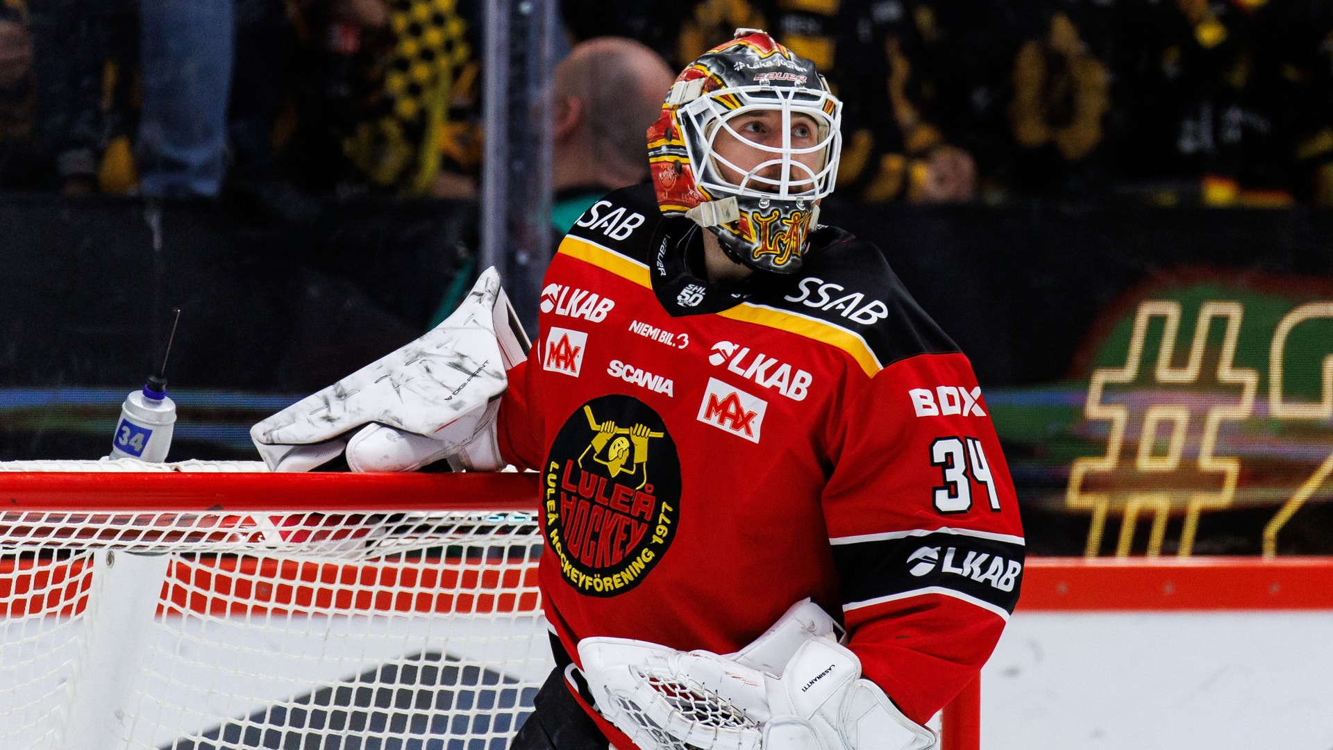 Ishockeyspelare i vit, gul och grön matchdräkt på isen under en match. Andra spelare och publik syns suddig i bakgrunden.