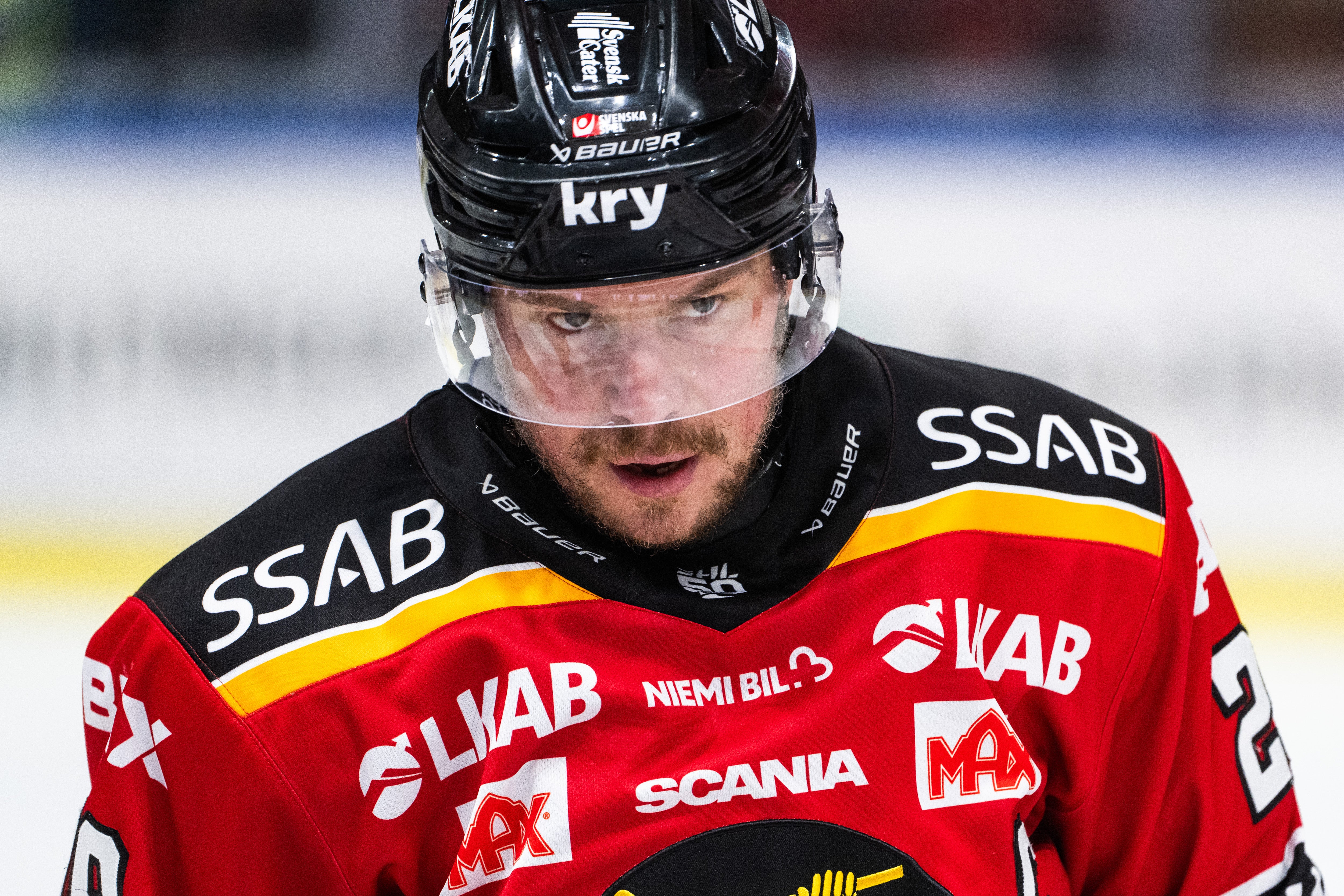 Ishockeyspelare i röd och svart tröja med hjälm och ansiktsskydd under en ishockeymatch.
