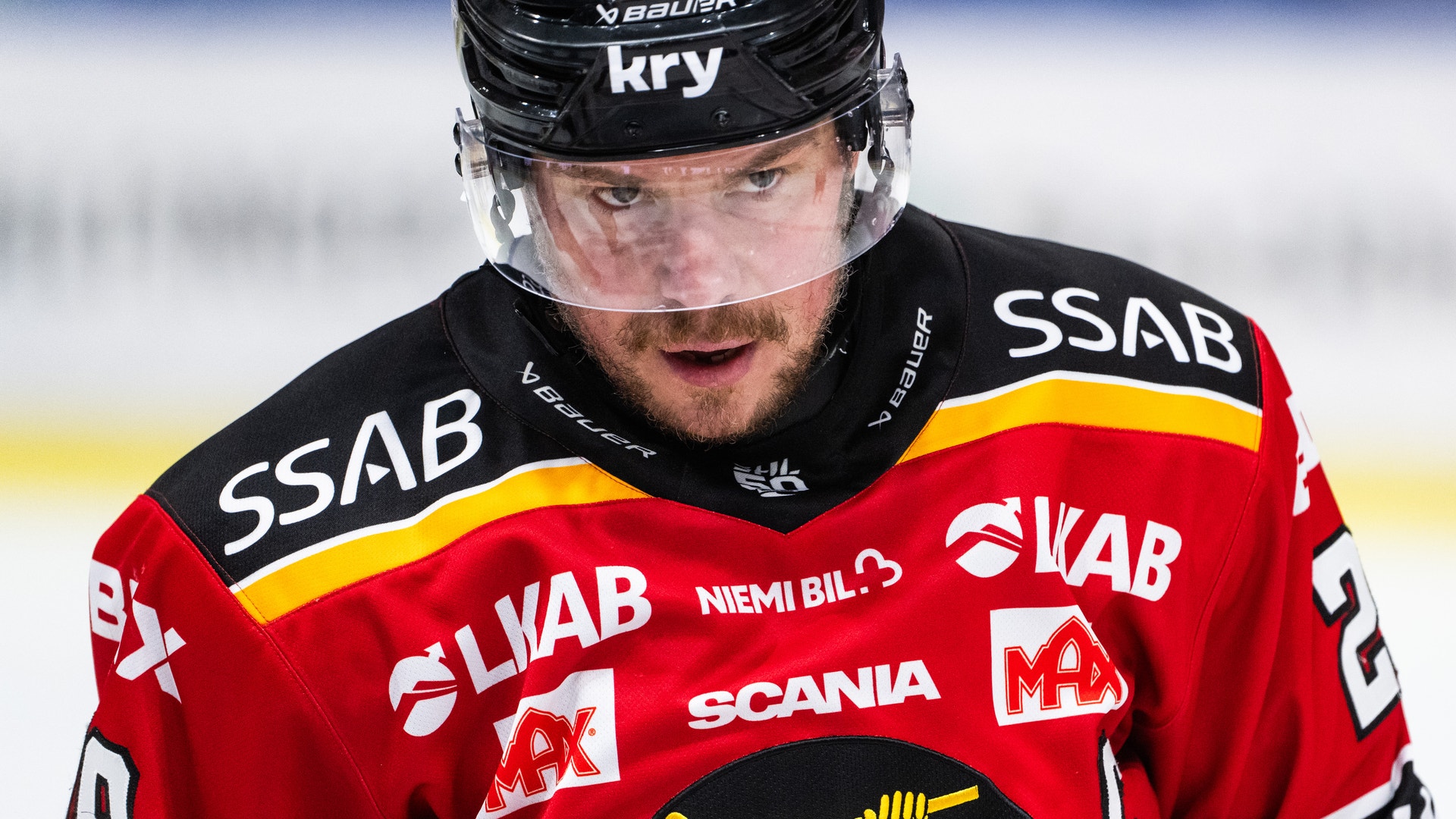 Ishockeyspelare i röd och svart tröja med hjälm och ansiktsskydd under en ishockeymatch.