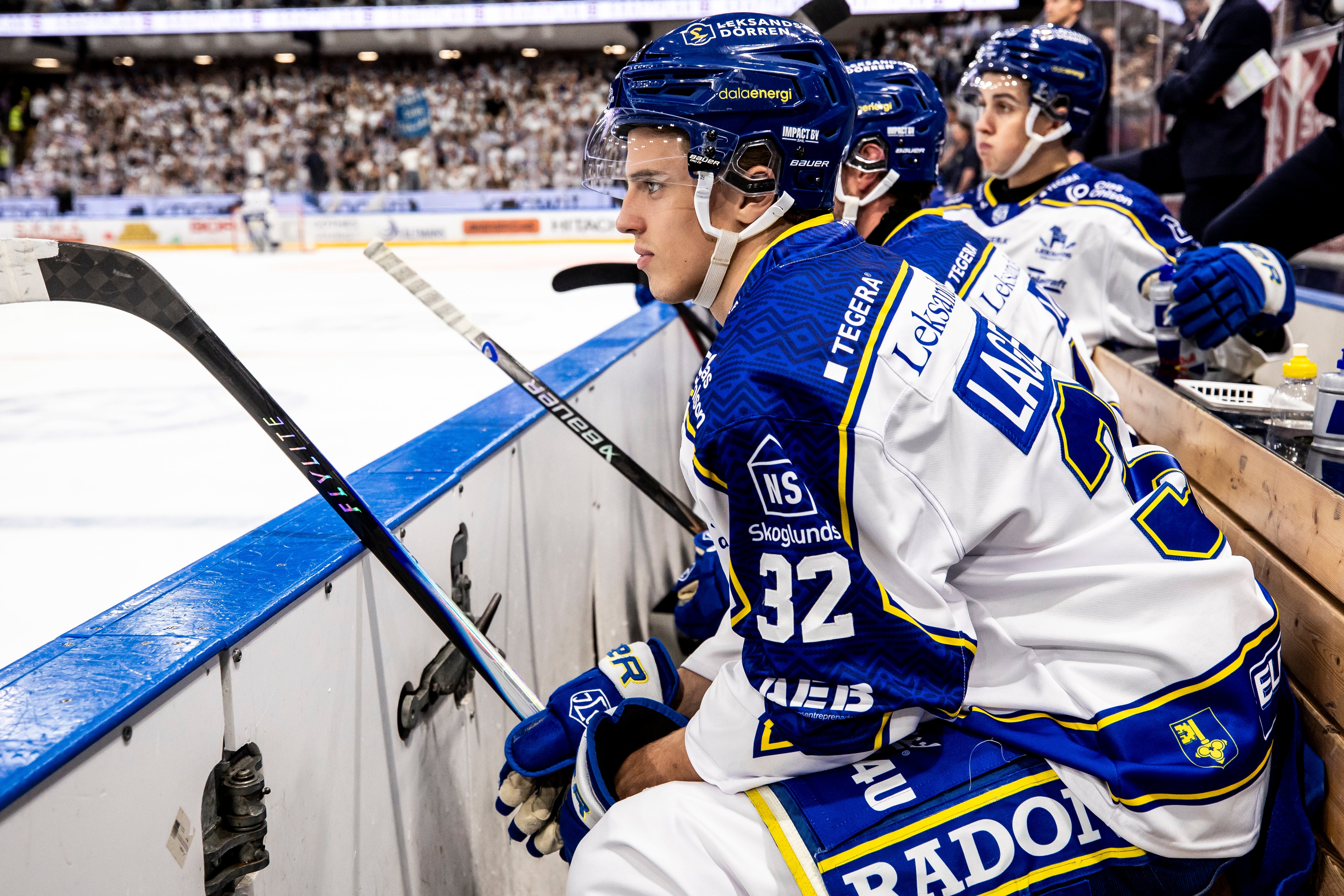 Ishockeyspelare i blå och vita trikåer sitter på bänken under en match i en fullsatt arena.