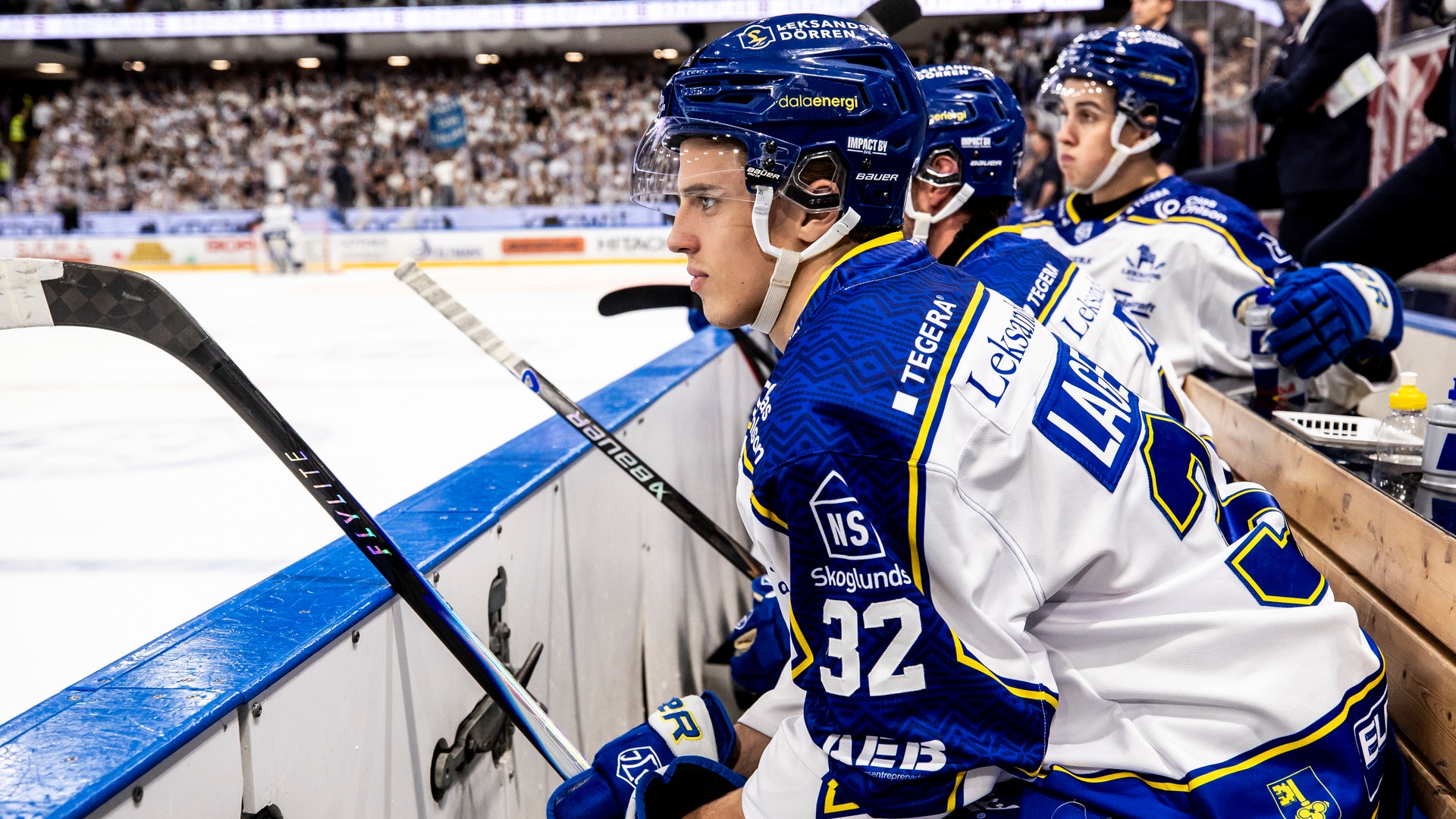 Ishockeyspelare i blå och vita trikåer sitter på bänken under en match i en fullsatt arena.