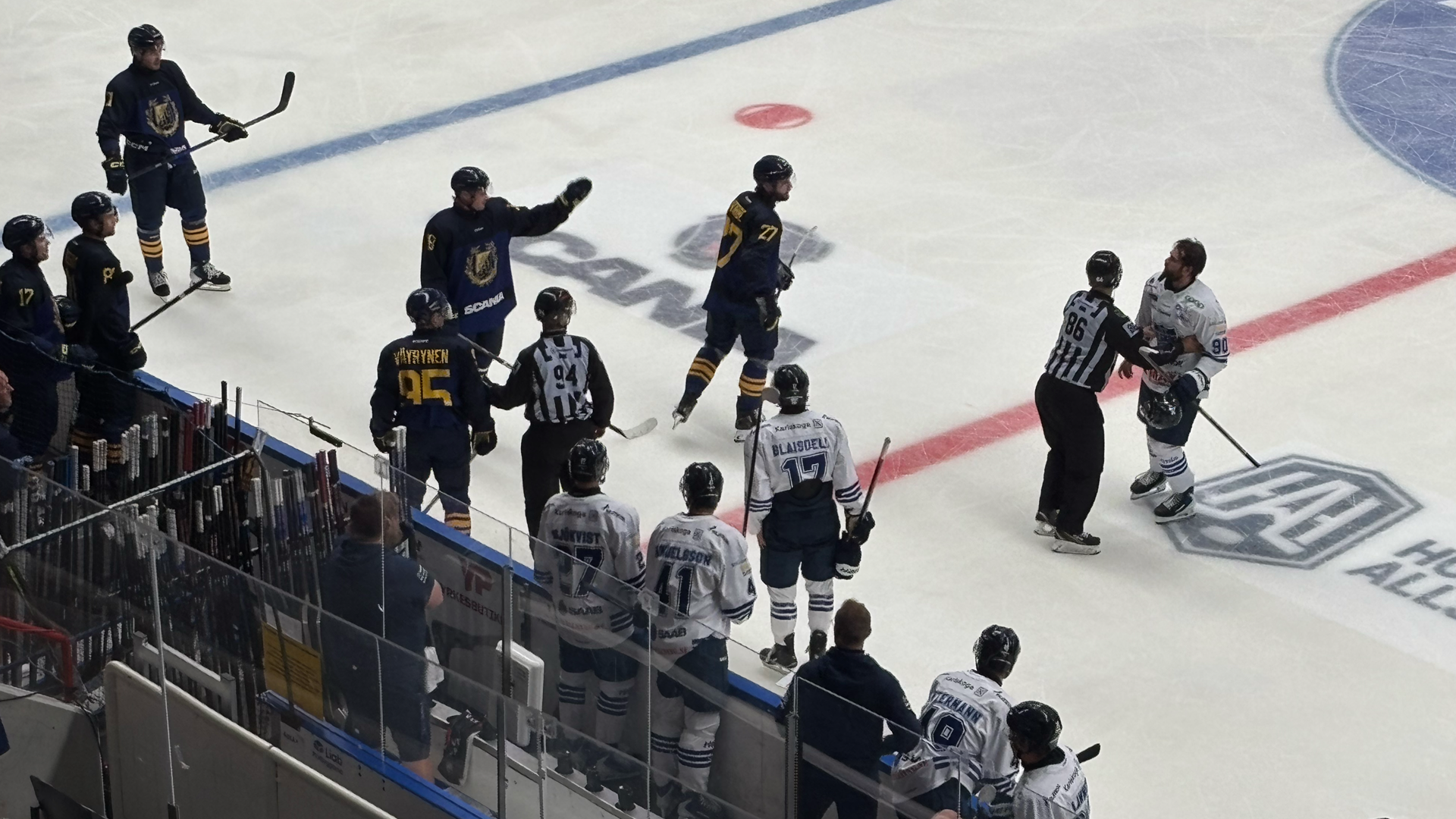 Översiktsbild från en ishockeymatch där två lag spelar på isen. Spelarna är klädda i svarta och vita trikåer och är utspridda på isbanan med en puck i spel. Bänkar med spelare och personal syns längs sidlinjen.