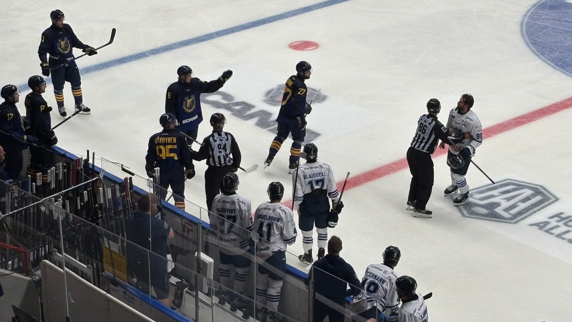 Översiktsbild från en ishockeymatch där två lag spelar på isen. Spelarna är klädda i svarta och vita trikåer och är utspridda på isbanan med en puck i spel. Bänkar med spelare och personal syns längs sidlinjen.
