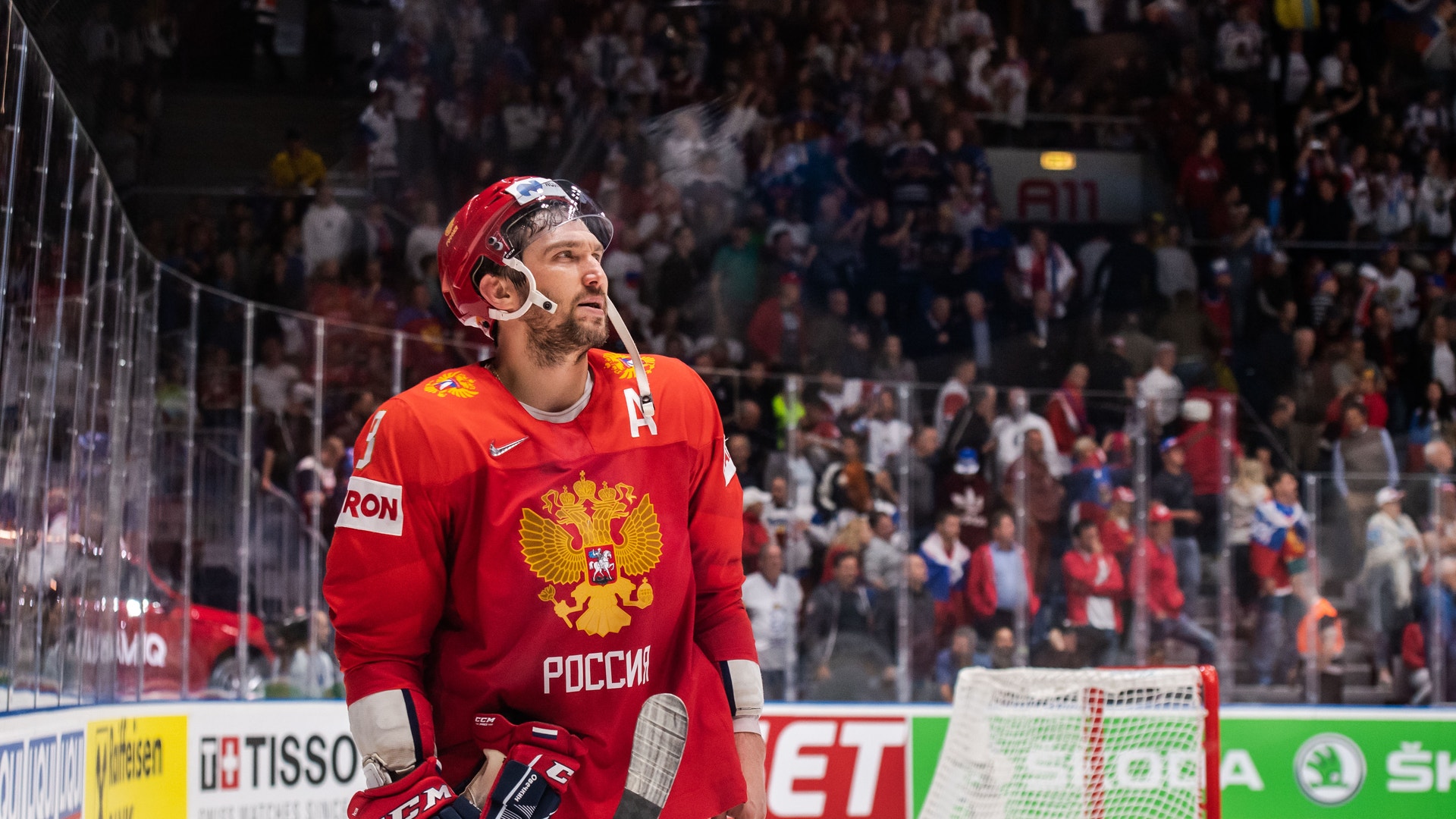 Hockeyspelar i röd landslagströja med rysk emblem står vid sargen under en ishockeymatch med fullsatt publik i bakgrunden.
