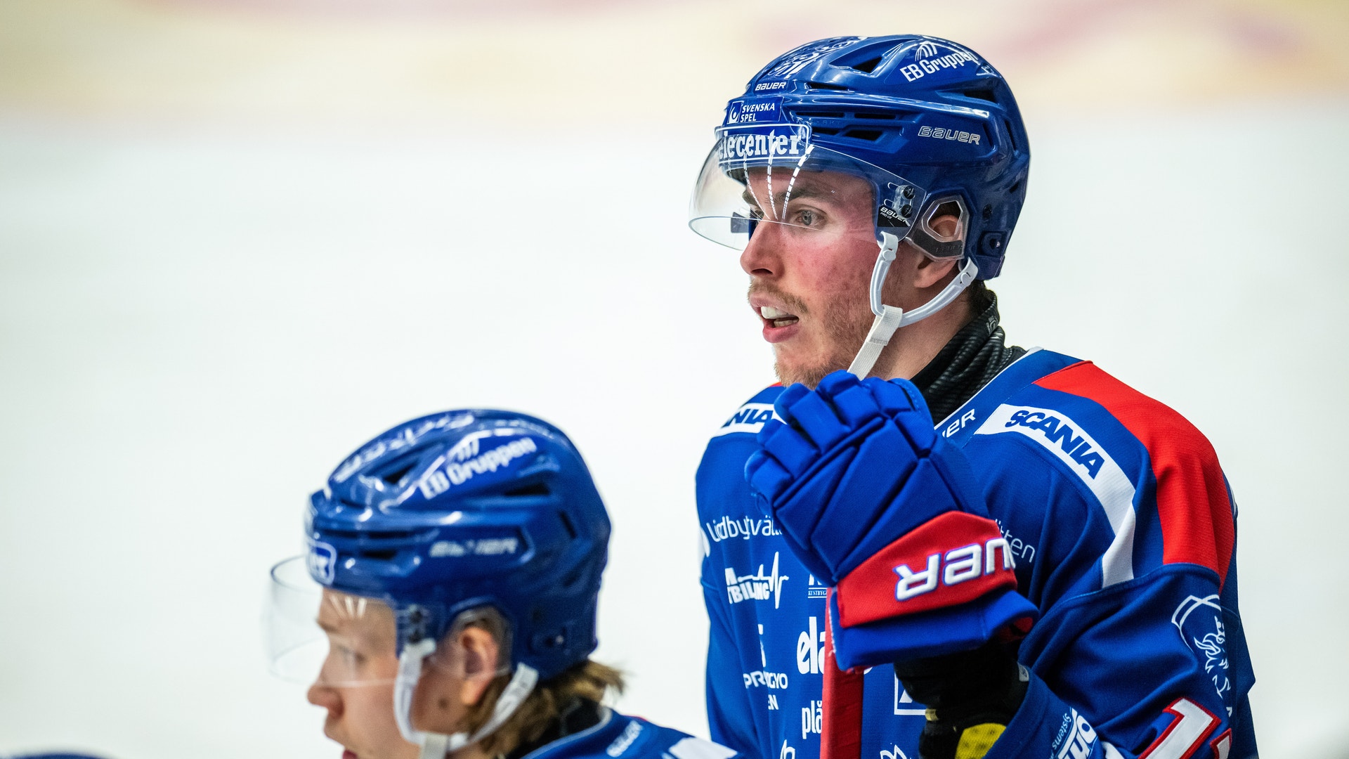 Två ishockeyspelare i blå och röda uniformer under en match. Båda bär skyddshjälmar och ser fokuserade ut under spelet.