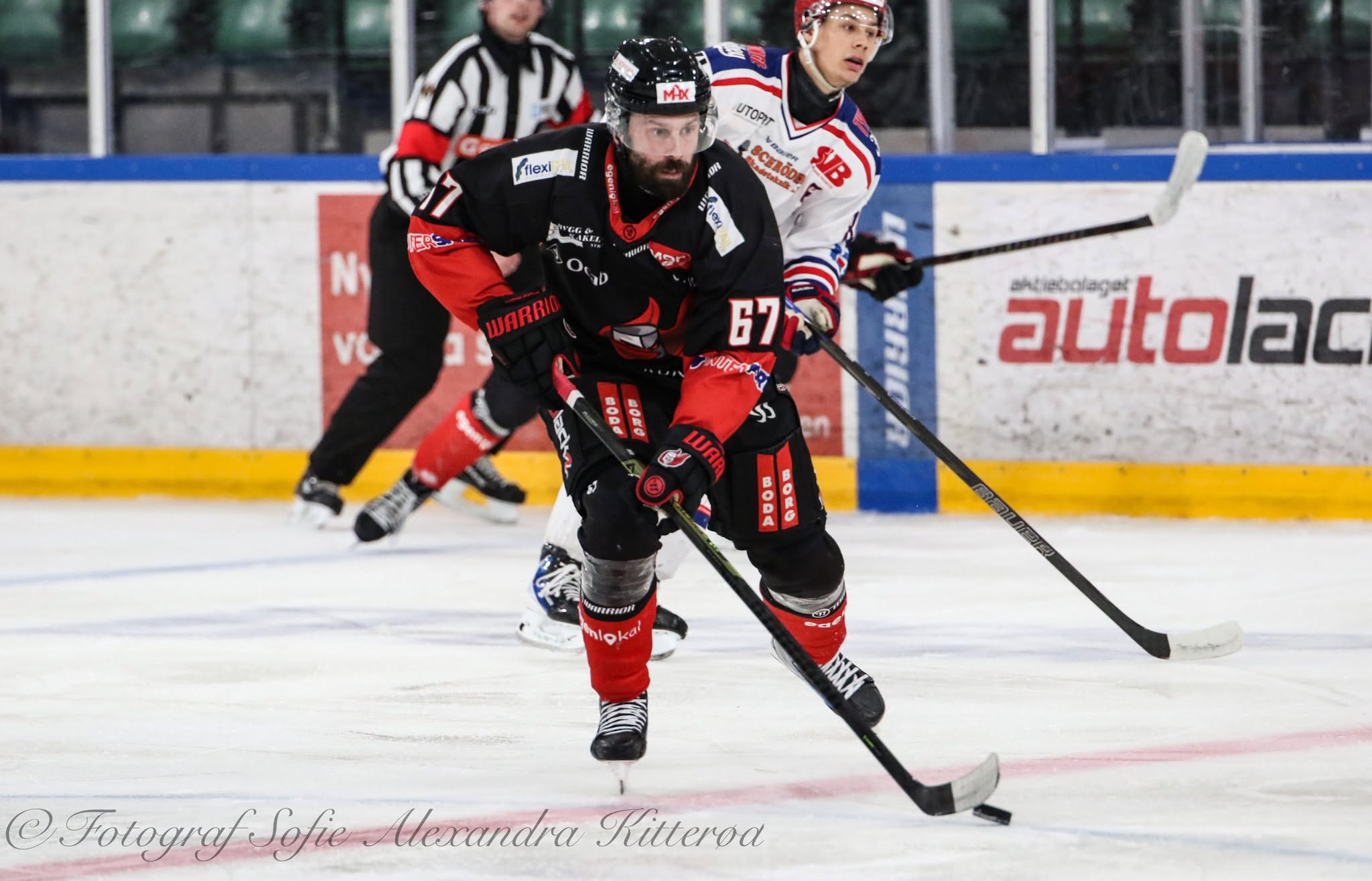 En ishockeyspelare i svart och rött matchställ med nummer 67 åker med pucken under en match. Bakom honom syns en motspelare i vit och blå tröja samt en domare.