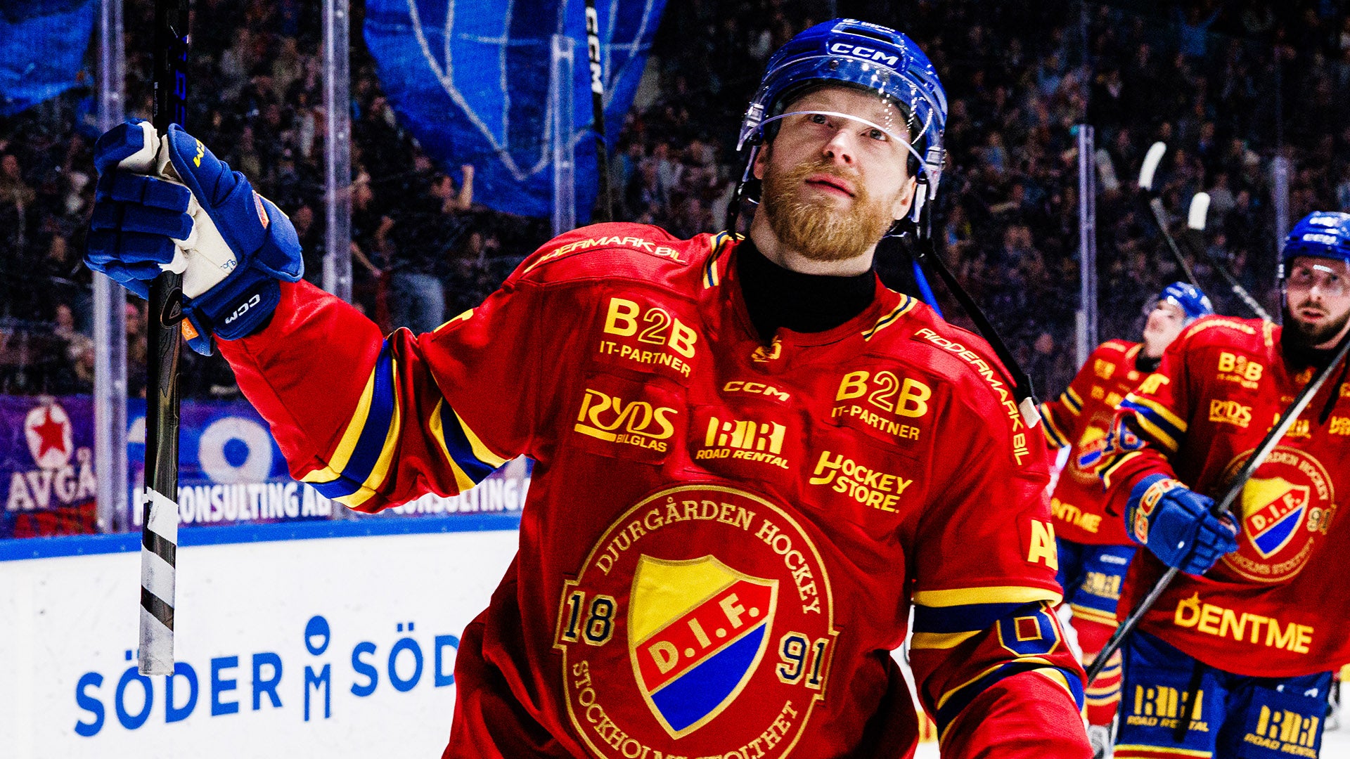 Ishockeyspelare i röd landslagströja jublar med höjd arm under en match. Spelaren bär hjälm och är omgiven av publik och lagkamrater i bakgrunden.