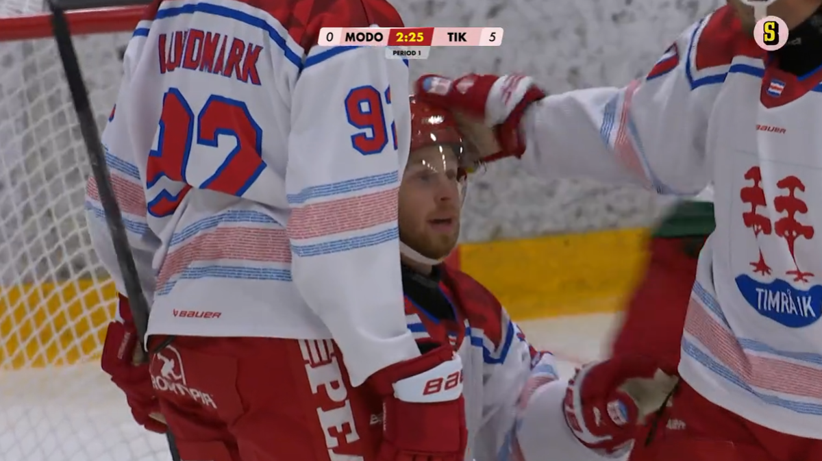 Ishockeyspelare i vita och röda trikåer samlas vid målet under en match. En spelare i röd tröja är nära mållinjen medan lagkamrater i vita trikåer är i närheten. Matchen visas på en tv-skärm med poängställning synlig.