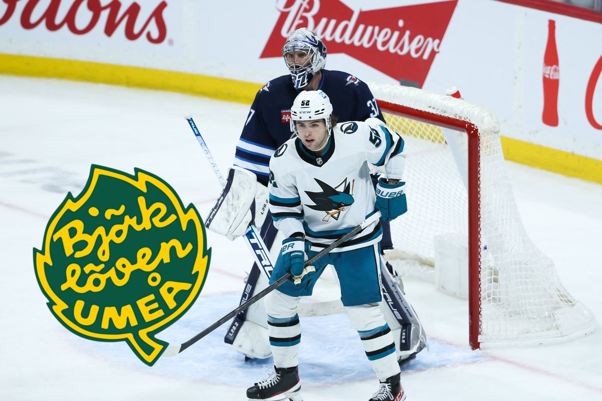 Ishockeymatch där en spelare i vit och blå tröja är positionerad framför målet med en försvarare i mörk tröja bakom honom. Publiken och sponsorloggor syns i bakgrunden.