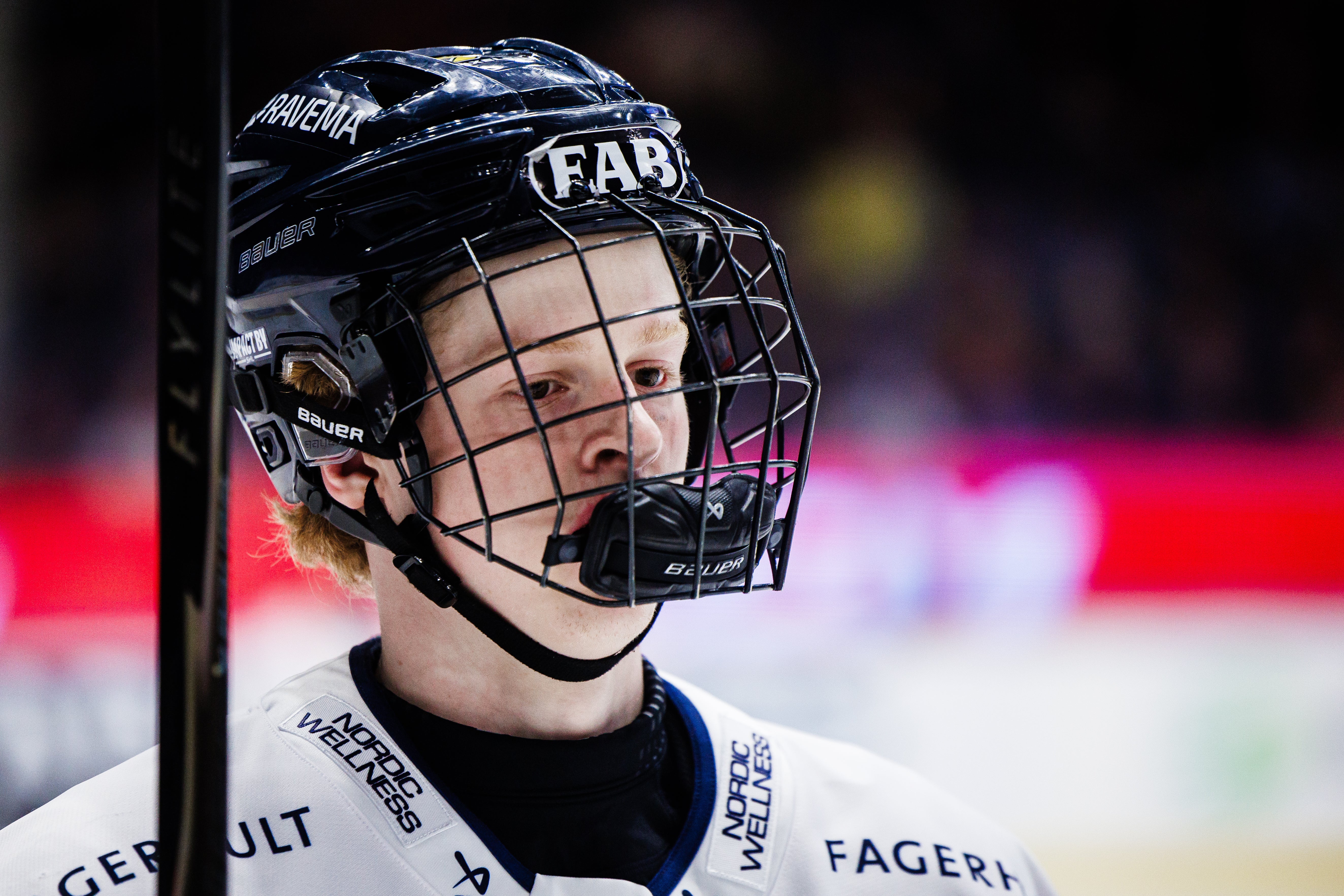 Ishockeyspelare i svart hjälm och vit tröja under en match. Spelaren tittar fokuserat framåt medan han håller i en hockeyklubb.