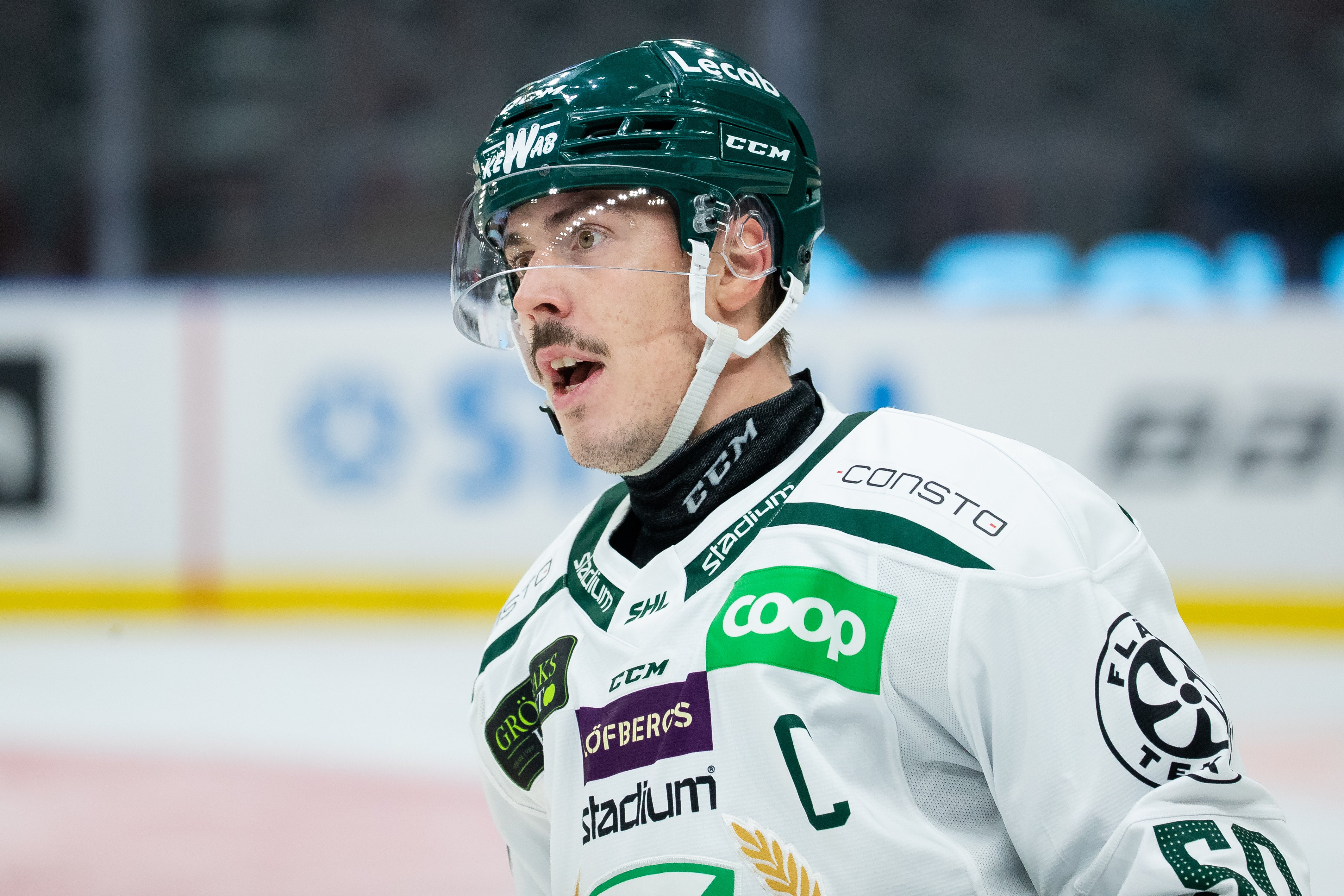 Ishockeyspelare i vit tröja med gröna detaljer under match. Spelaren bär grön hjälm och har ett fokuserat uttryck.