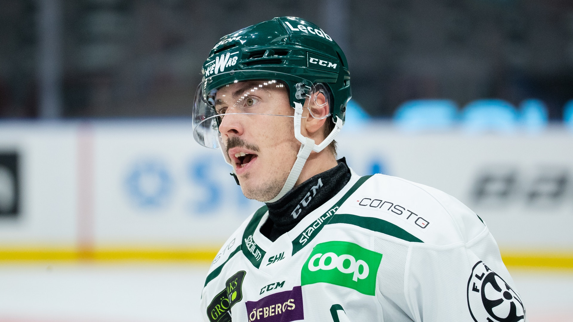 Ishockeyspelare i vit tröja med gröna detaljer under match. Spelaren bär grön hjälm och har ett fokuserat uttryck.