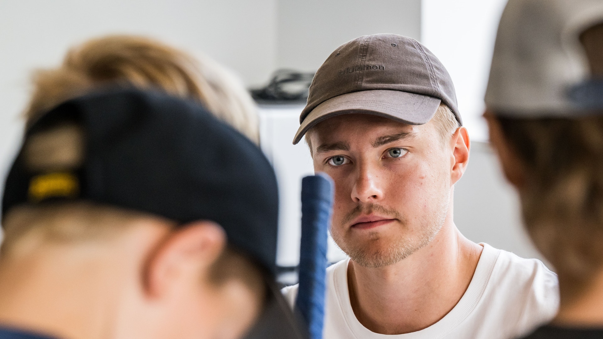 En man i vit t-shirt och grå keps står tillsammans med andra personer i vad som verkar vara en sportmiljö.