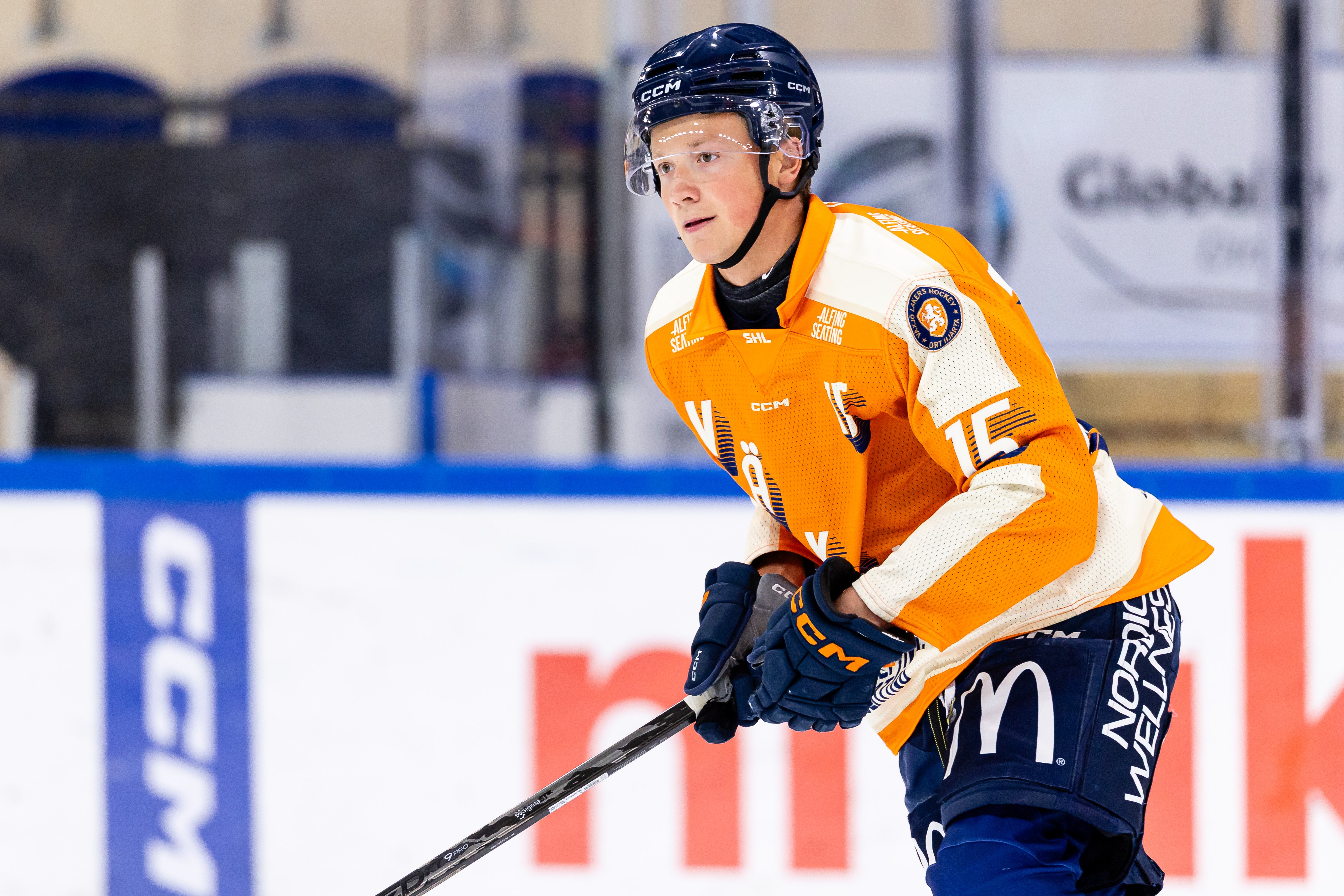 En ung ishockeyspelare i orange tröja och mörkblå hjälm åker på isen med klubban i händerna.