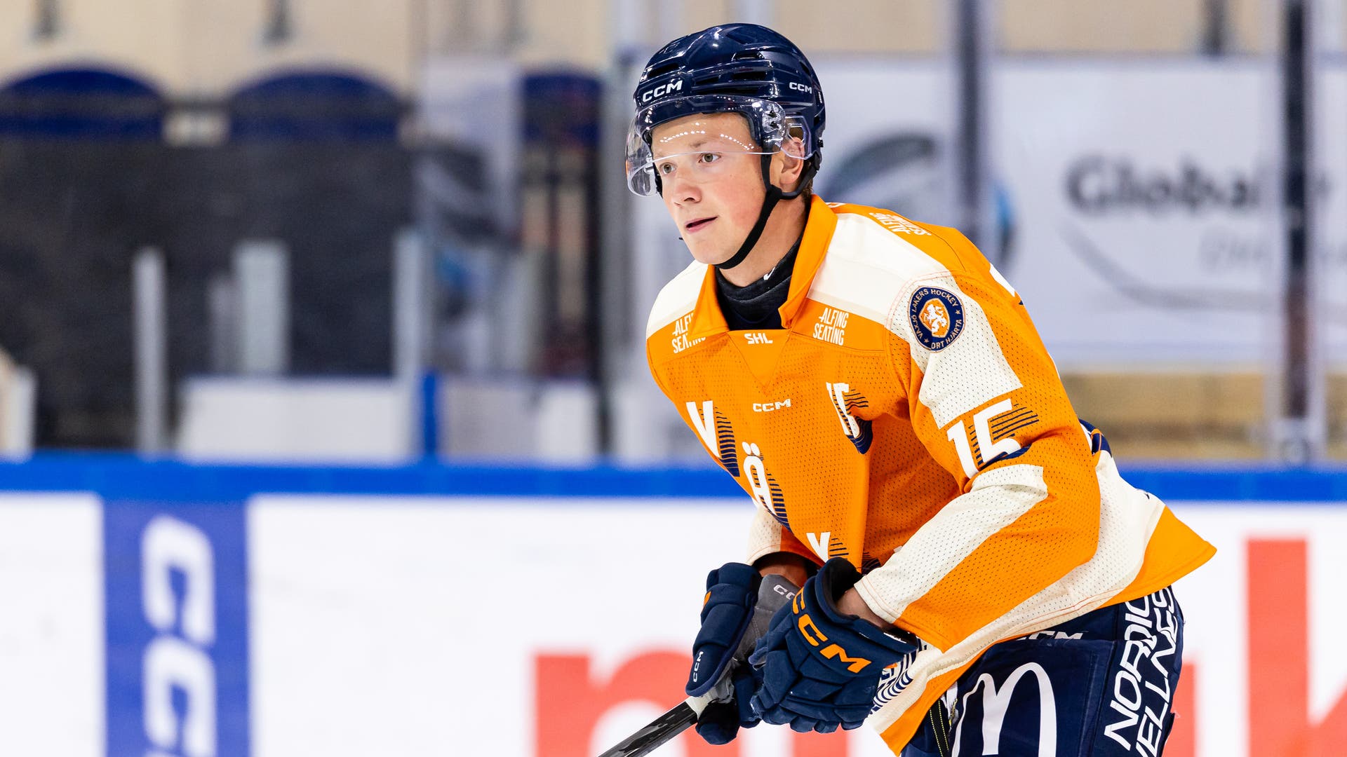 En ung ishockeyspelare i orange tröja och mörkblå hjälm åker på isen med klubban i händerna.