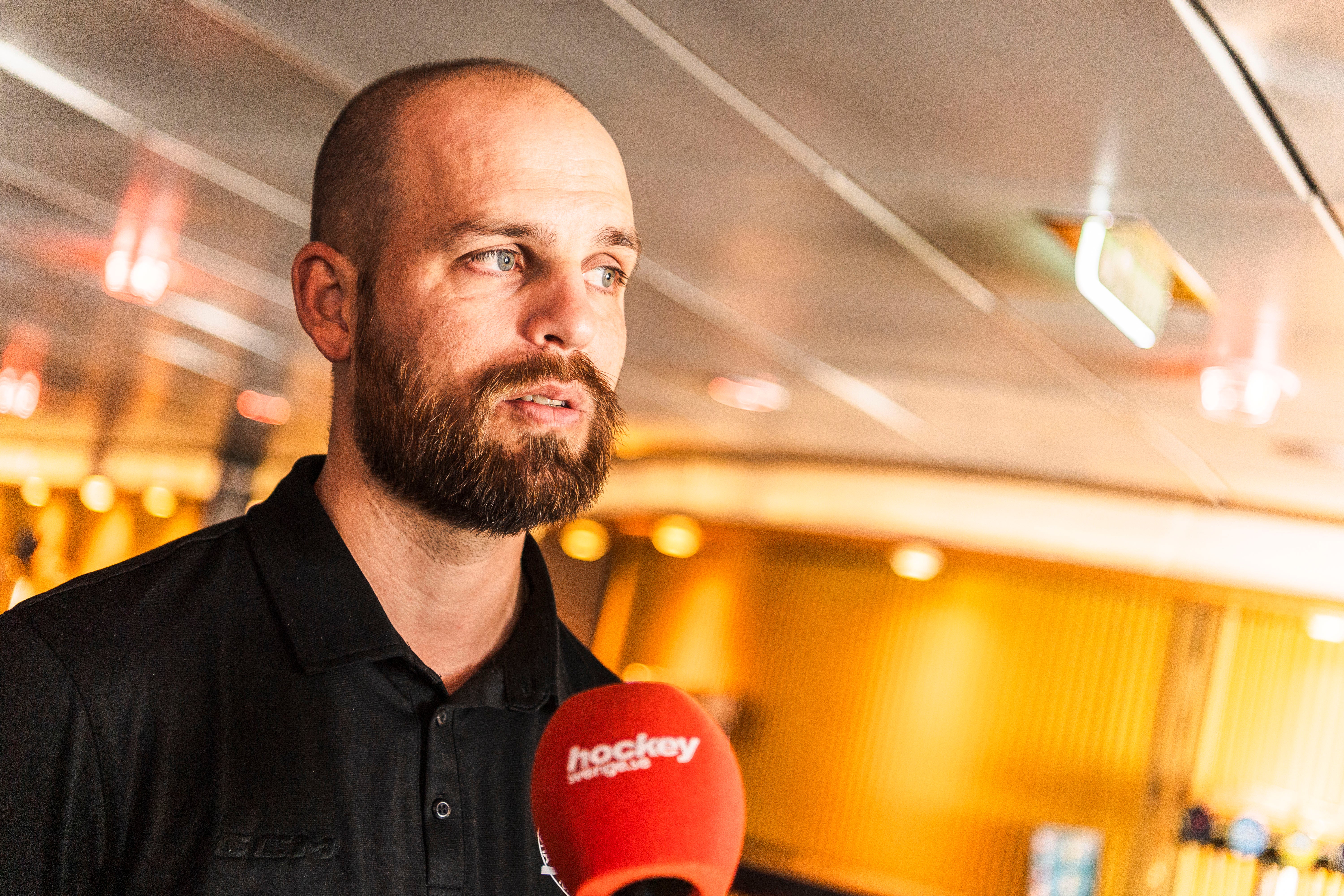 Porträtt av en man med skägg som håller en röd mikrofon under en intervju eller presskonferens. Han är klädd i svart och ser ut mot sidan. Bakgrunden visar varma gula ljus som skapar en professionell miljö.