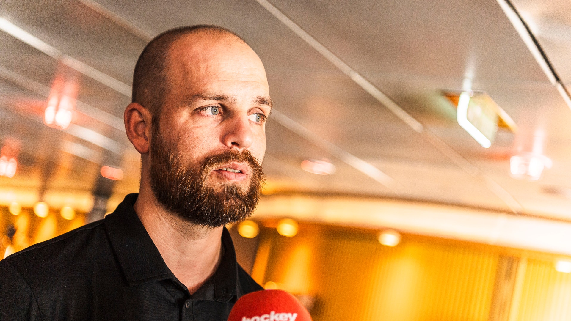 Porträtt av en man med skägg som håller en röd mikrofon under en intervju eller presskonferens. Han är klädd i svart och ser ut mot sidan. Bakgrunden visar varma gula ljus som skapar en professionell miljö.