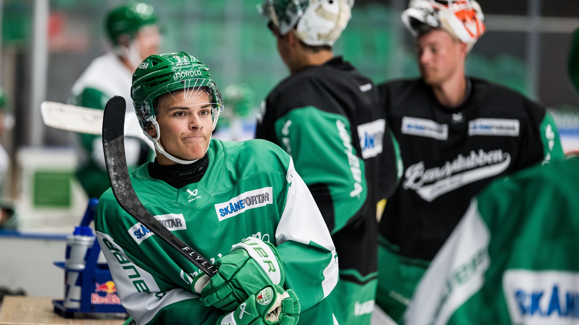 Ishockeyspelare i grön och vit matchdräkt står på isen med sin stick och handskar. Andra spelare och tränare syns i bakgrunden.