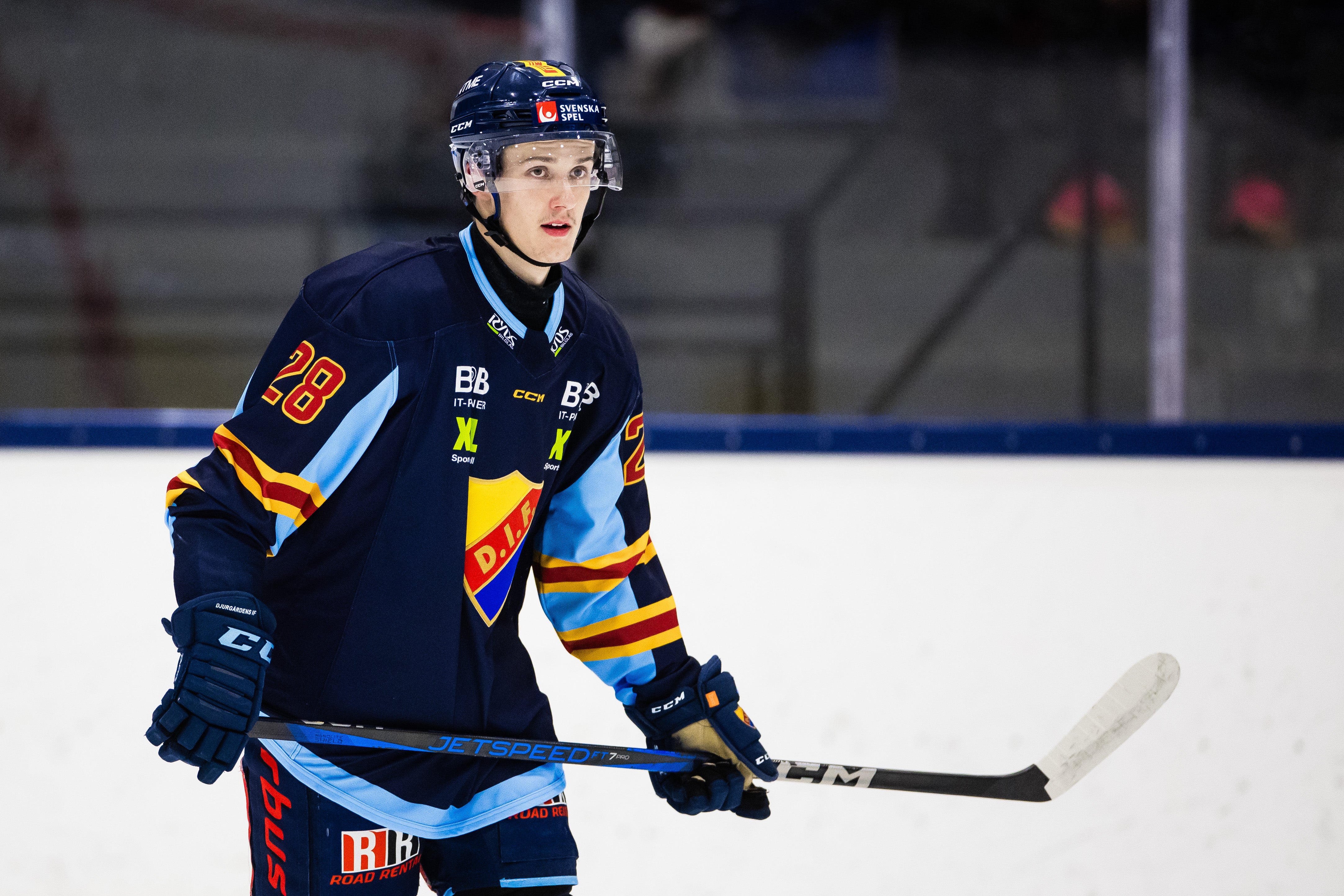 Ishockeyspelare i mörkblå lag-tröja med nummer 28, stående på isen med hockeyklubb i händerna under en match.