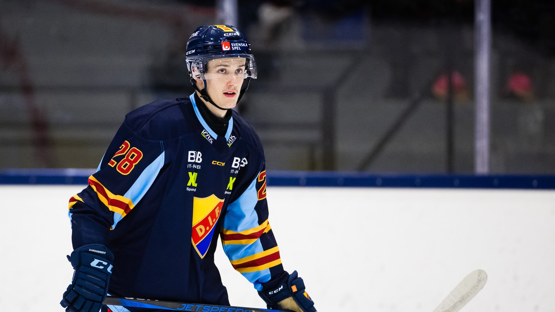 Ishockeyspelare i mörkblå lag-tröja med nummer 28, stående på isen med hockeyklubb i händerna under en match.