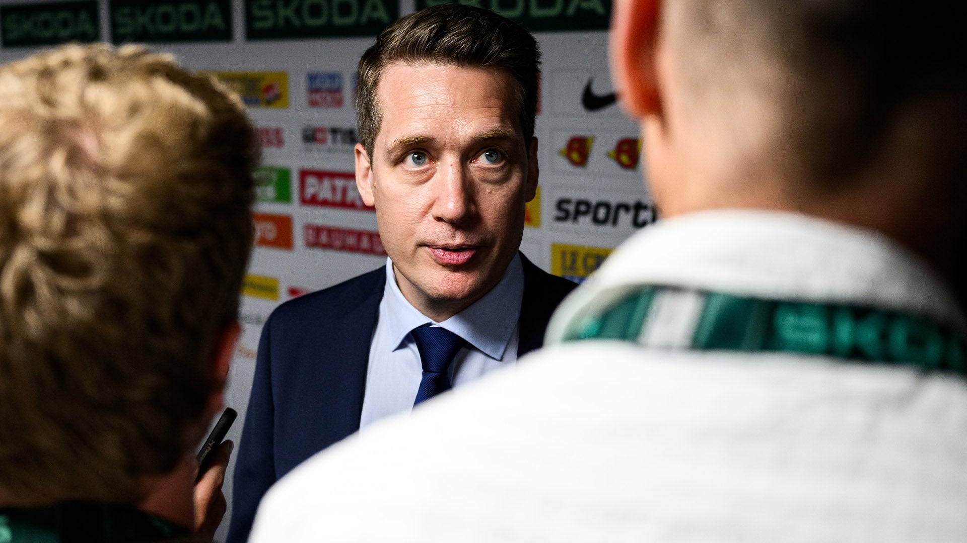 Man i mörkblå kostym och slips under intervju vid en sportrelaterad händelse med sponsorlogotyper i bakgrunden.