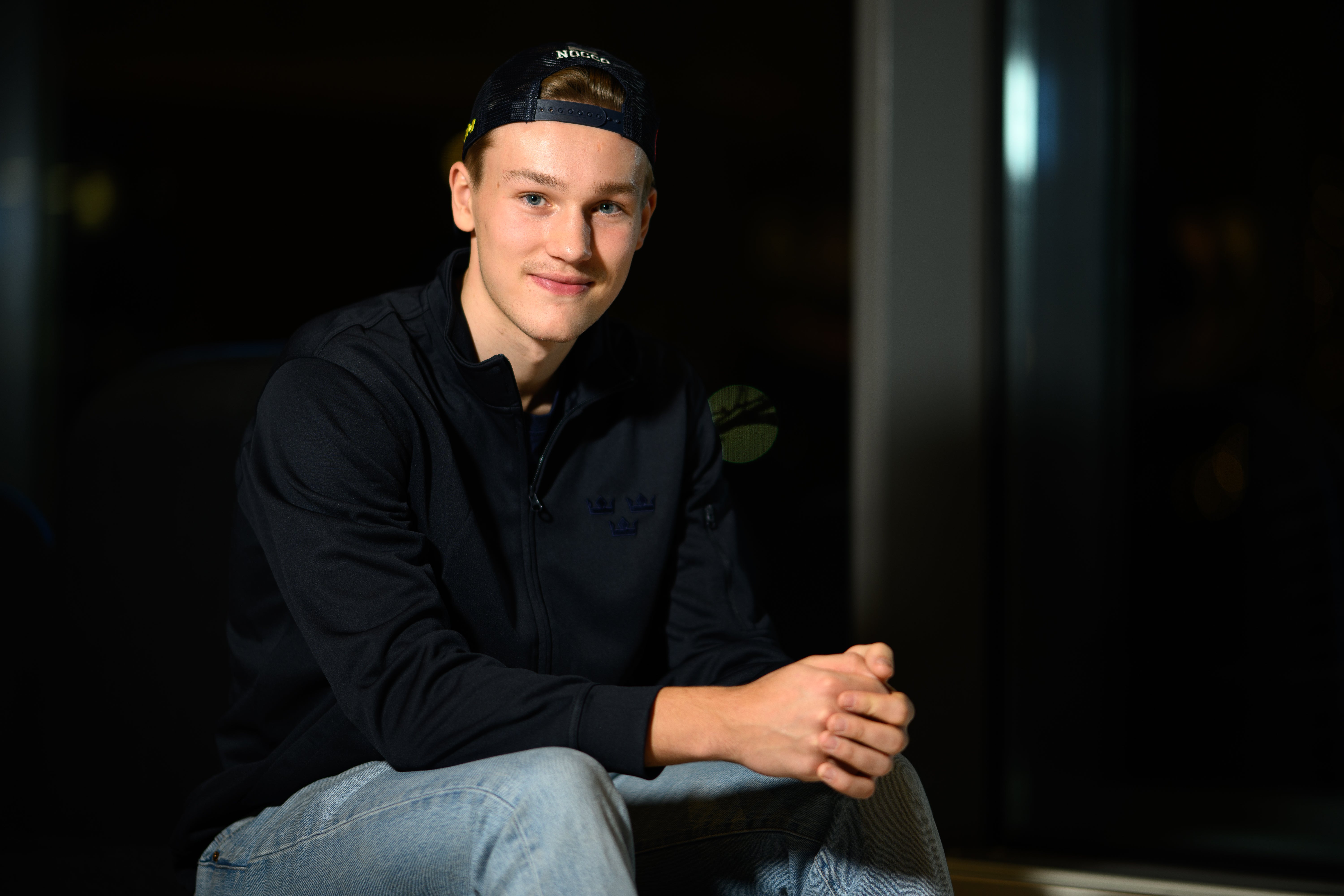 Porträtt av en ung man som sitter och poserar för kameran. Han bär en svart jacka och ljusa jeans med en keps på huvudet.