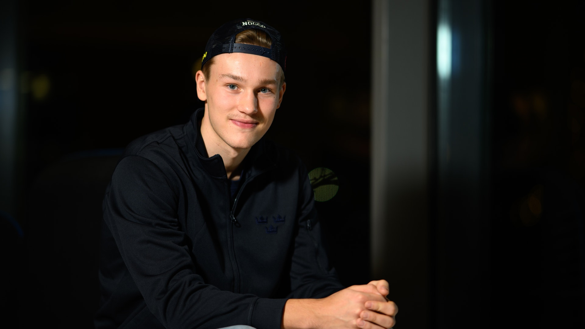 Porträtt av en ung man som sitter och poserar för kameran. Han bär en svart jacka och ljusa jeans med en keps på huvudet.