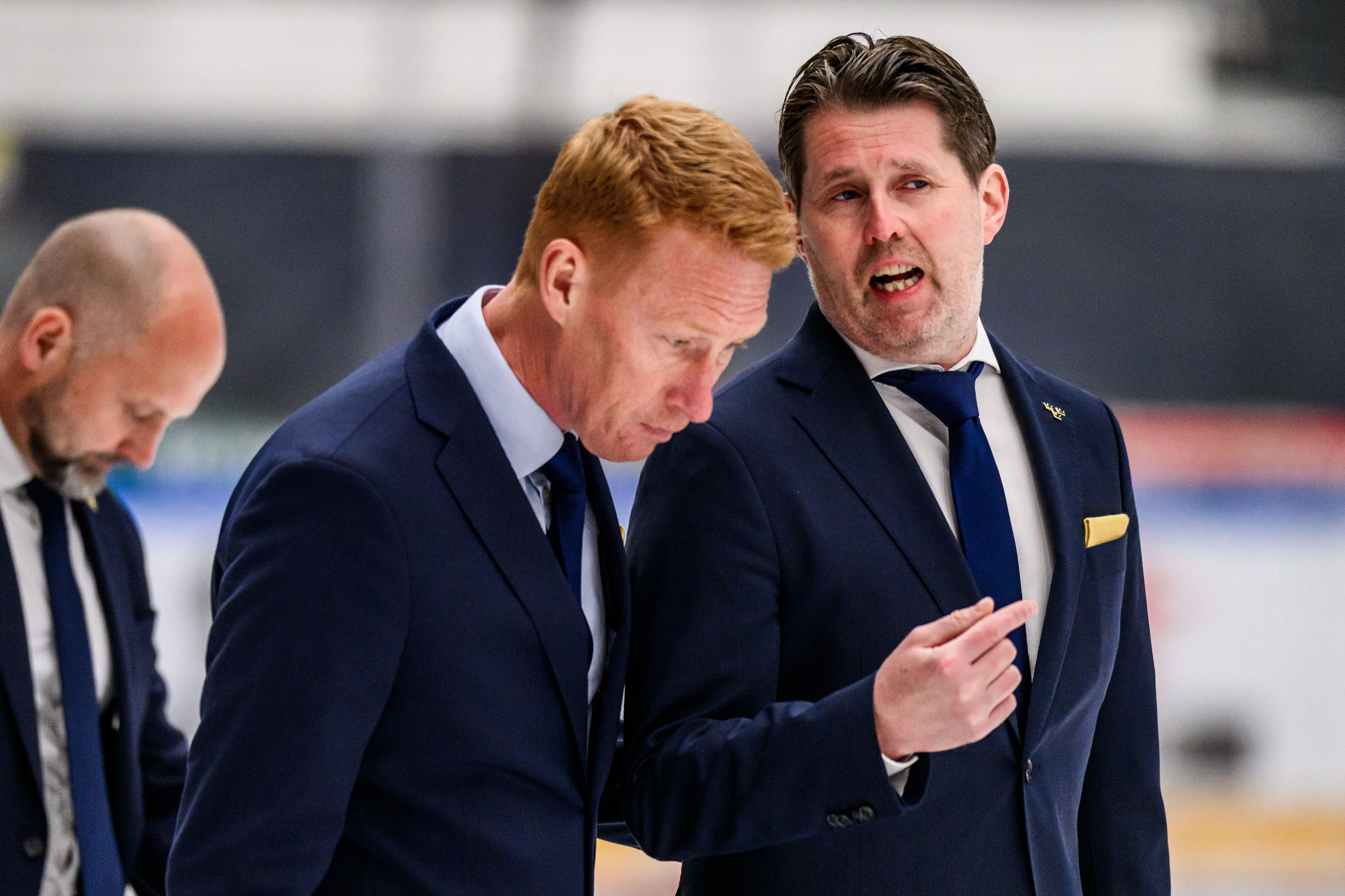 Tre män i mörka kostymer diskuterar på en ishockeyrink.