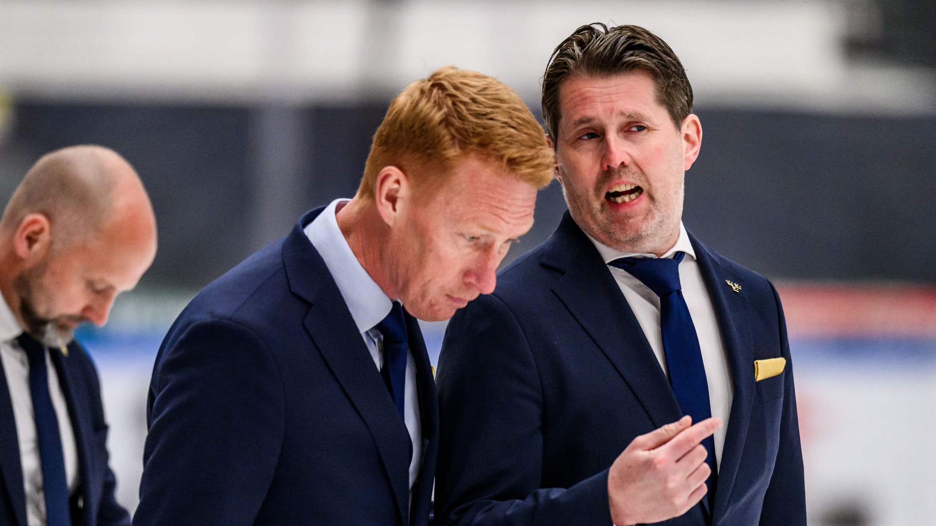 Tre män i mörka kostymer diskuterar på en ishockeyrink.