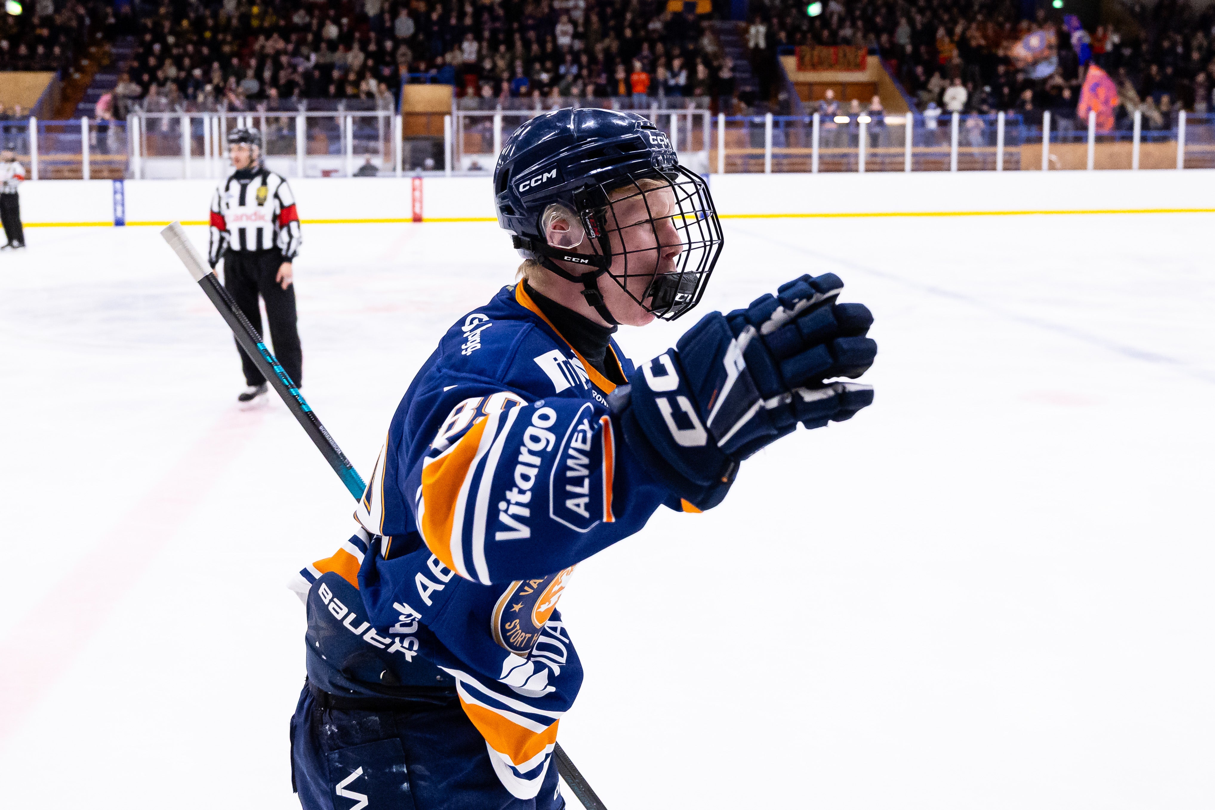 En ishockeyspelare i blå och orange matchdräkt firar under en match på isen. Spelaren bär hjälm och handskar, med en domare och publik synlig i bakgrunden.