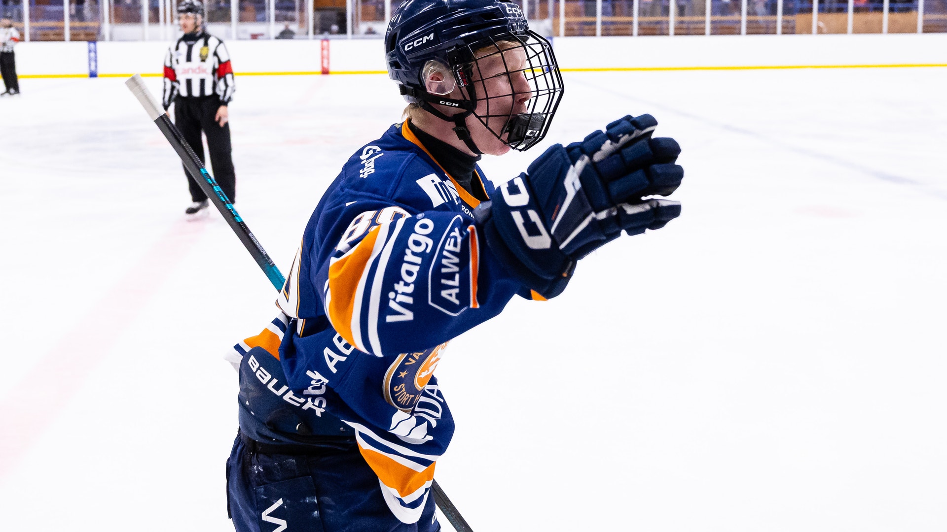 En ishockeyspelare i blå och orange matchdräkt firar under en match på isen. Spelaren bär hjälm och handskar, med en domare och publik synlig i bakgrunden.
