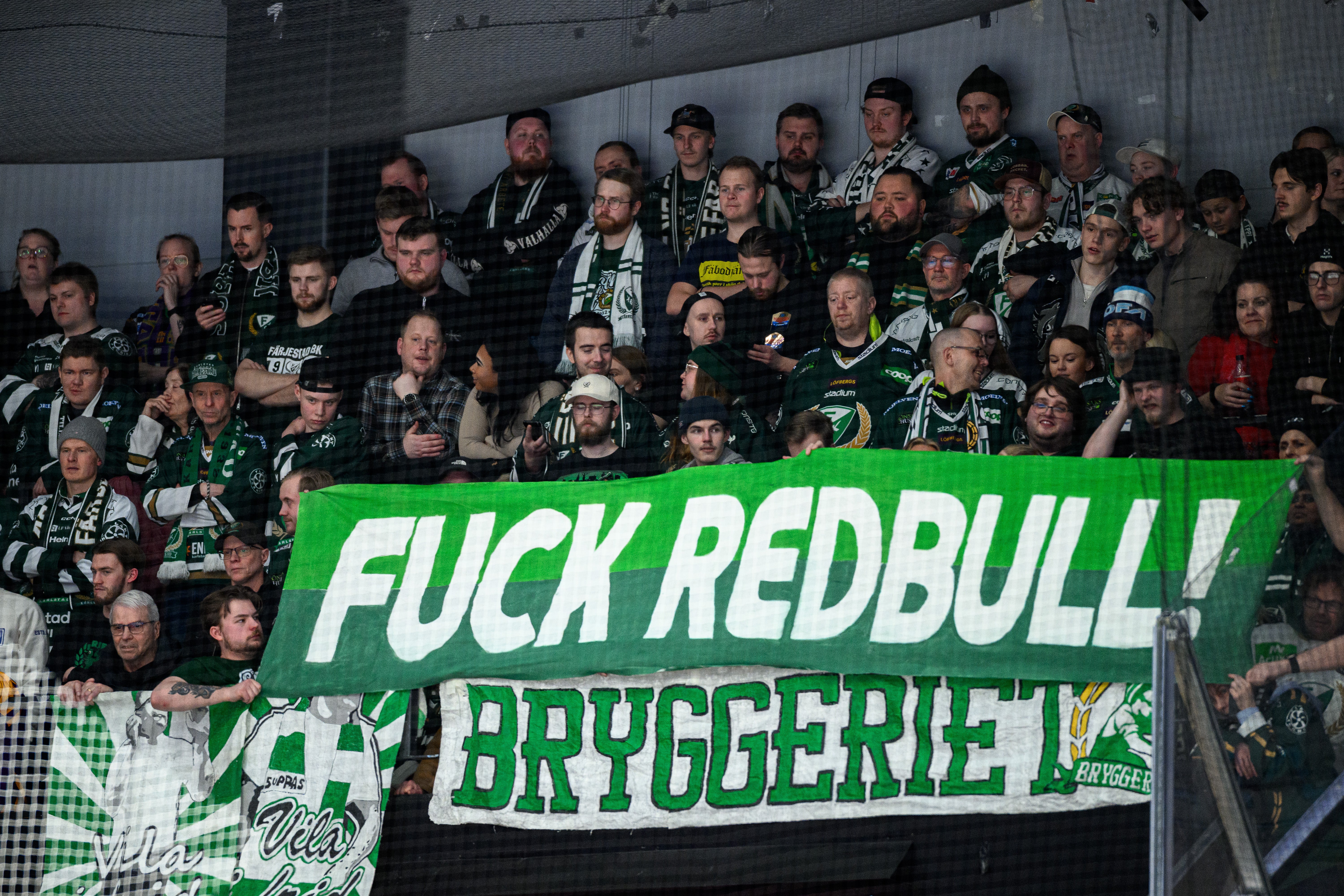 En grupp hockeysupportrar på läktaren håller upp en grön och vit banderoll med en provocerande slogan under en ishockeymatch.
