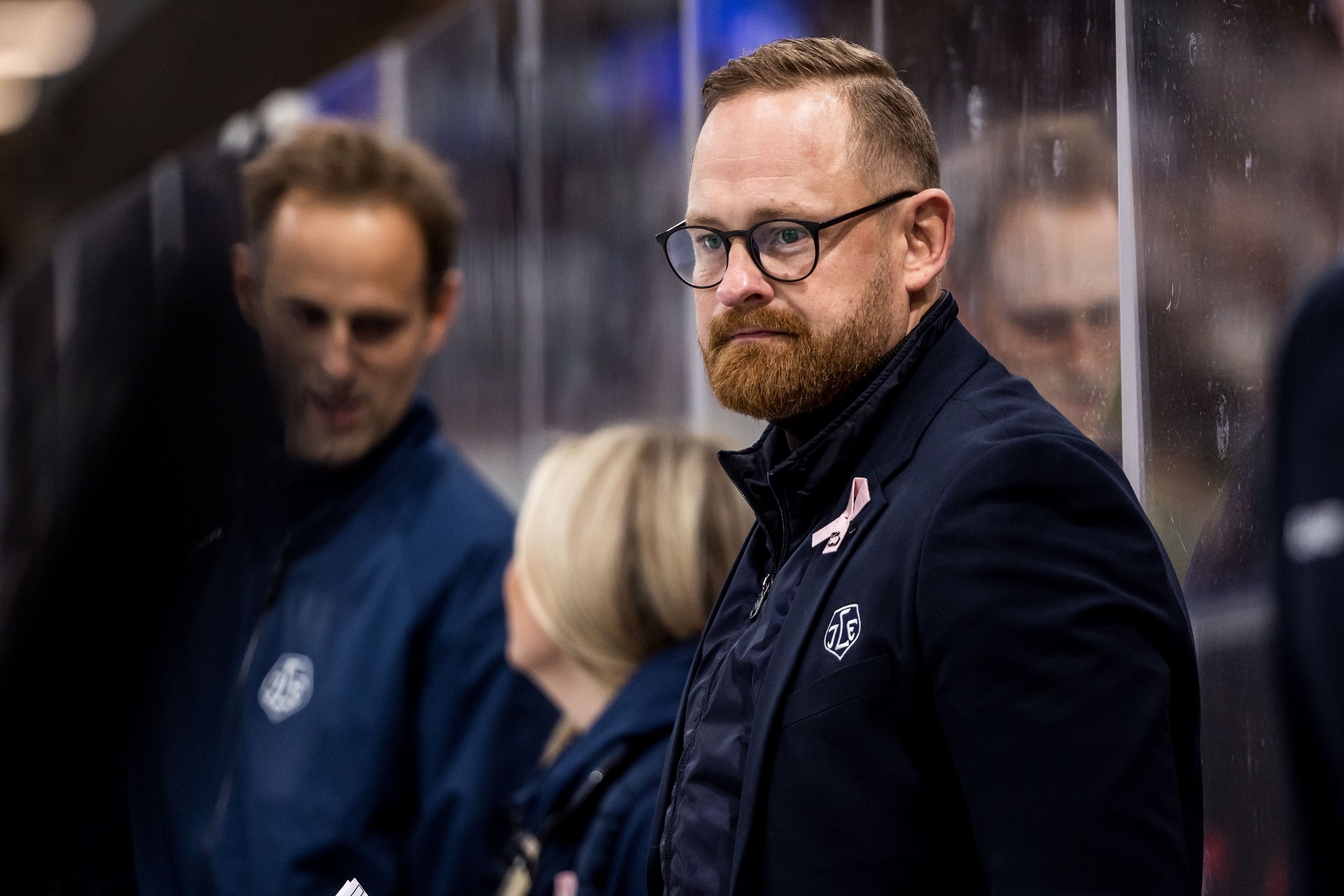 En man med rödbrunt skägg och glasögon, klädd i mörk jacka, står vid avbytarbänken under en ishockeymatch och tittar mot kameran. Bakom honom syns två personer i suddig bakgrund.